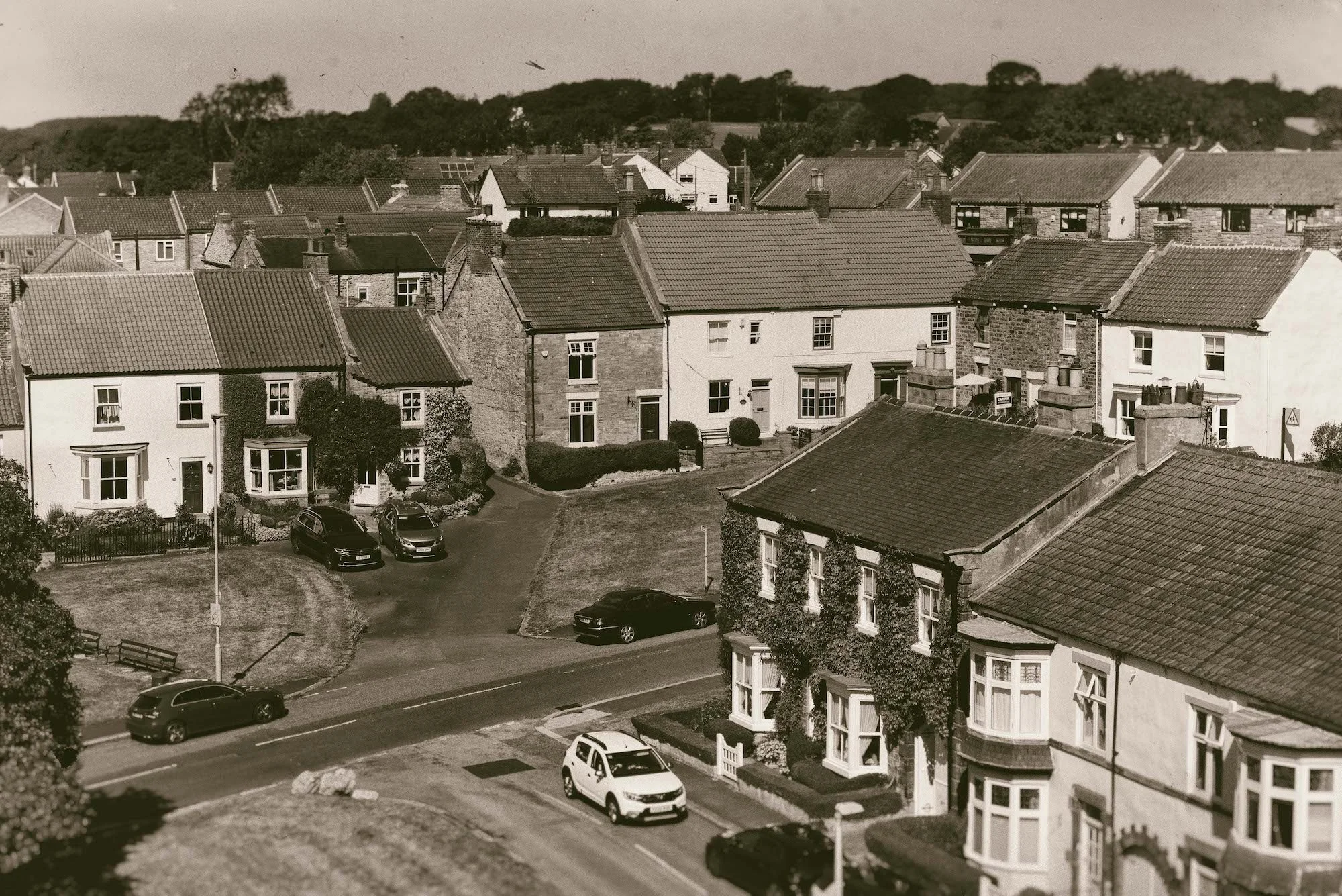 Black and white photo of a residential neighborhood with closely built houses, some with bay windows and chimneys, and a small parking area with cars. There are trees and hills in the background.