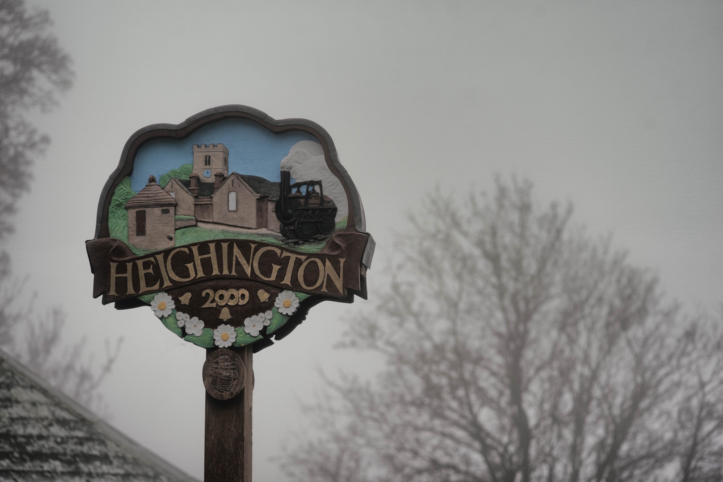 A decorative signpost with the town name Hegington, 2020, featuring a painted scene of a church with a clock tower, a small building, and a steam locomotive, surrounded by white daisies with yellow centers. The sign is mounted on a wooden post with a