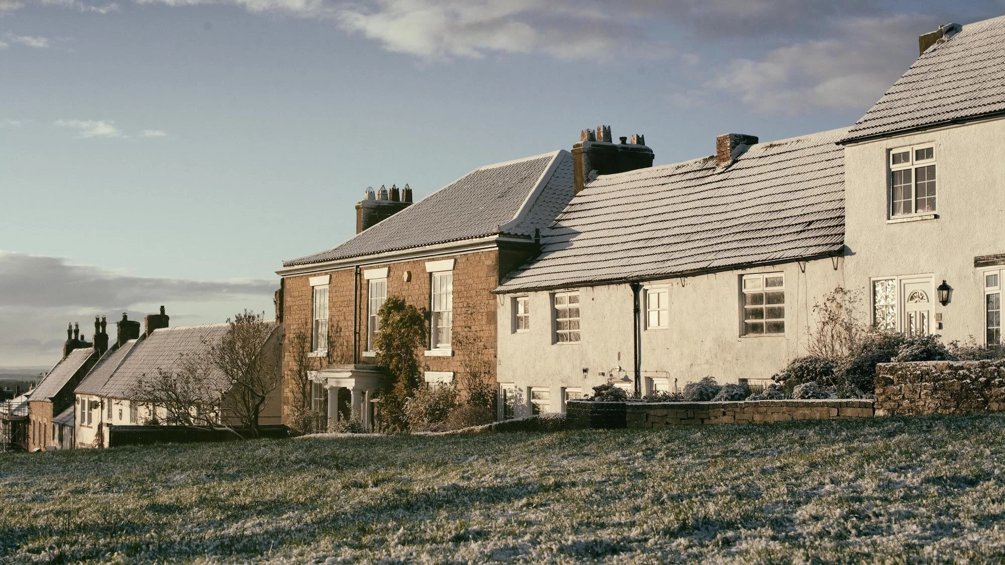 A row of houses with snow on the roofs, set against a clear sky, with a grassy foreground and trees.