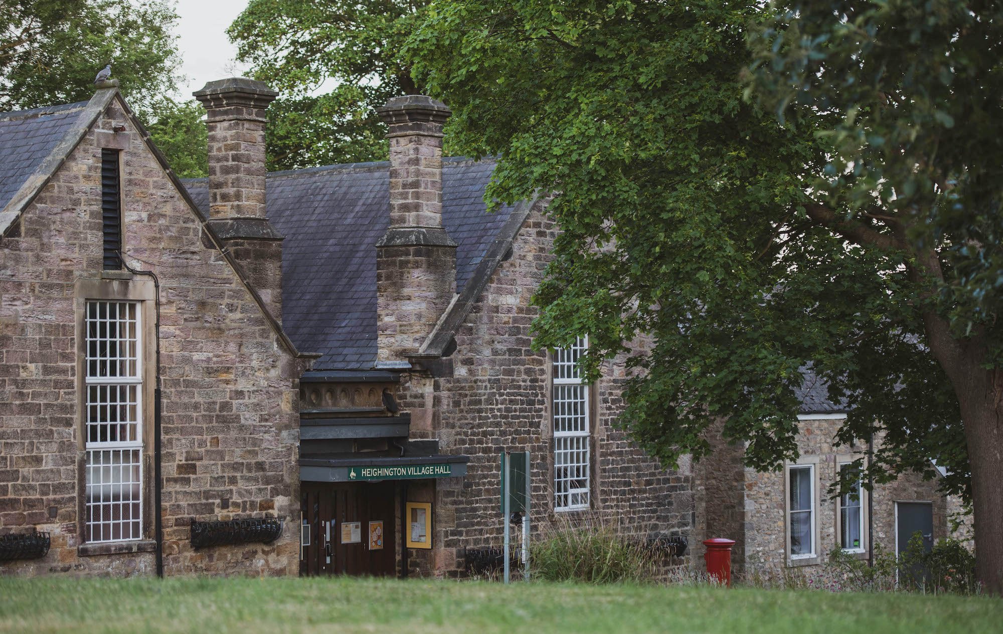 A stone building with a sign reading 'Heightington Village Hall' surrounded by trees and greenery.