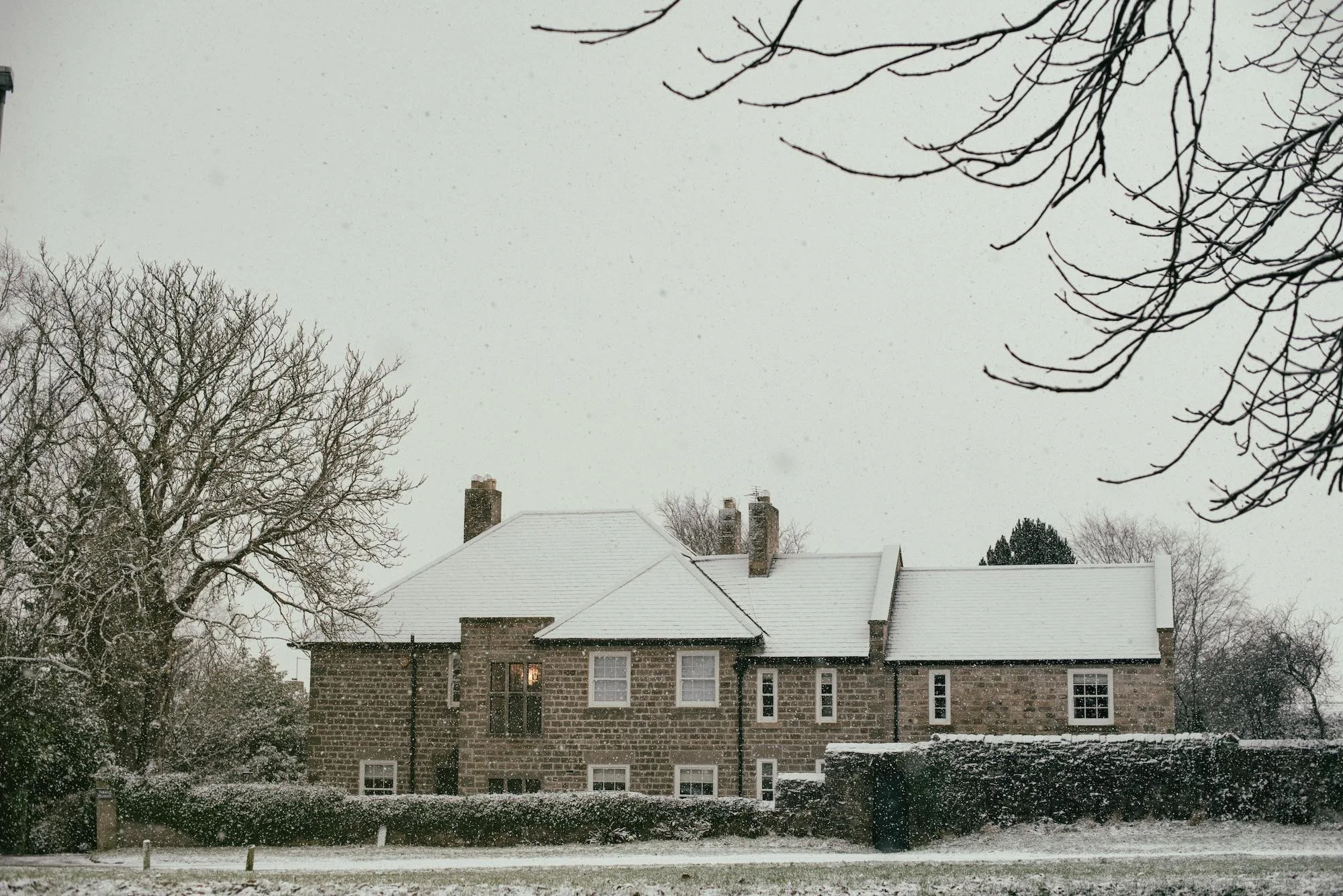 Snow-covered house with brick exterior and multiple chimneys, surrounded by snow-covered trees, under a gray cloudy sky during a snowy day.