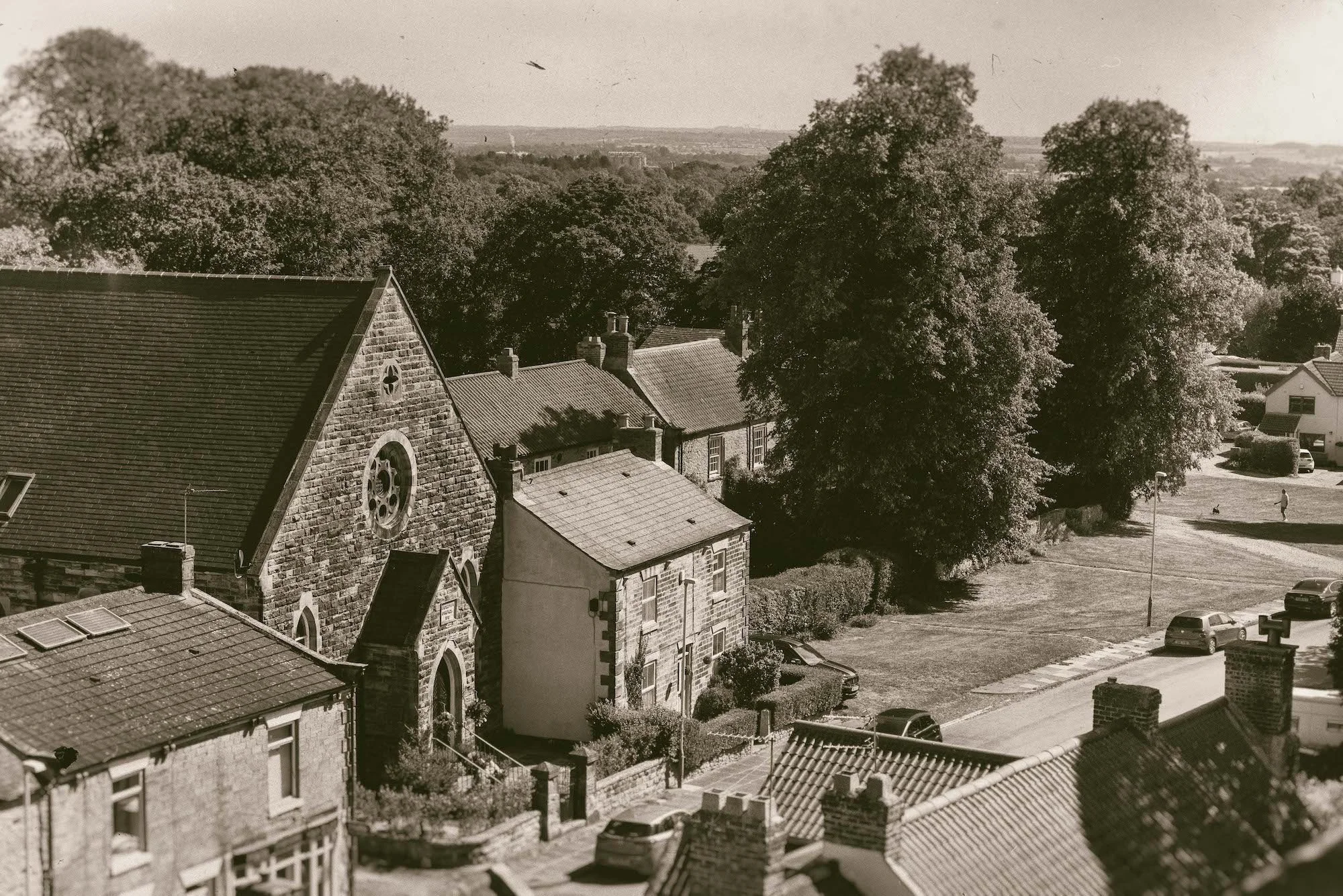 Black-and-white photo of a small town with stone buildings, large trees, a church with a pointed roof, parked cars, and a street.