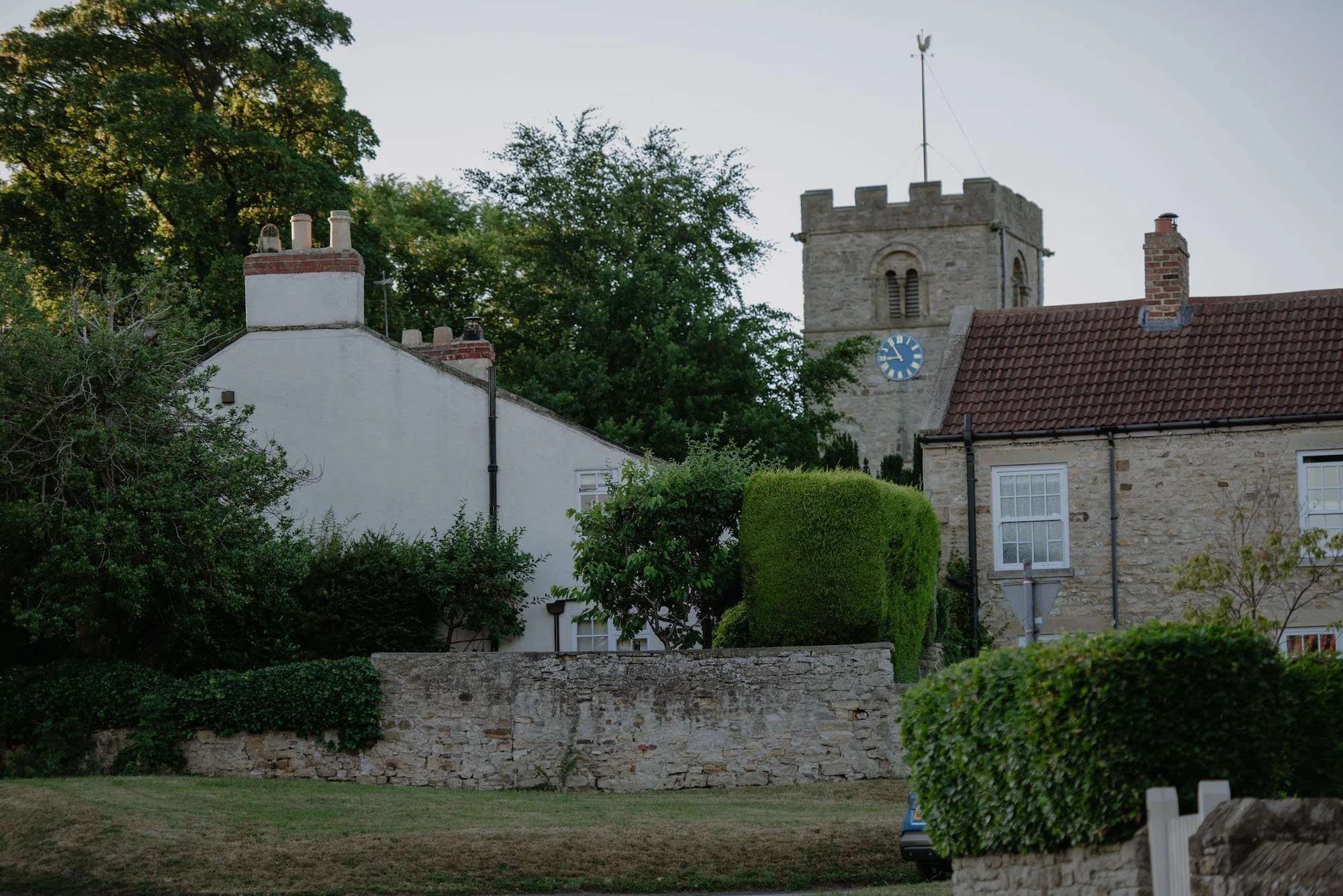 View of a historic stone church with a square tower featuring battlements, a blue clock face, and a weather vane on top, surrounded by residential houses with white facades, brick chimneys, and lush green trees and bushes in the foreground.