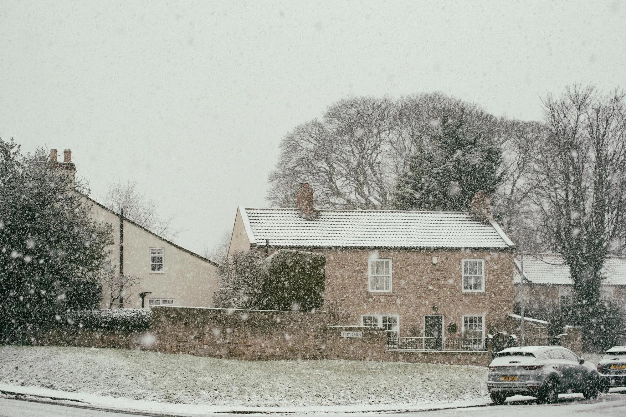 Snow falling over residential buildings with a stone wall in the foreground and trees in the background.