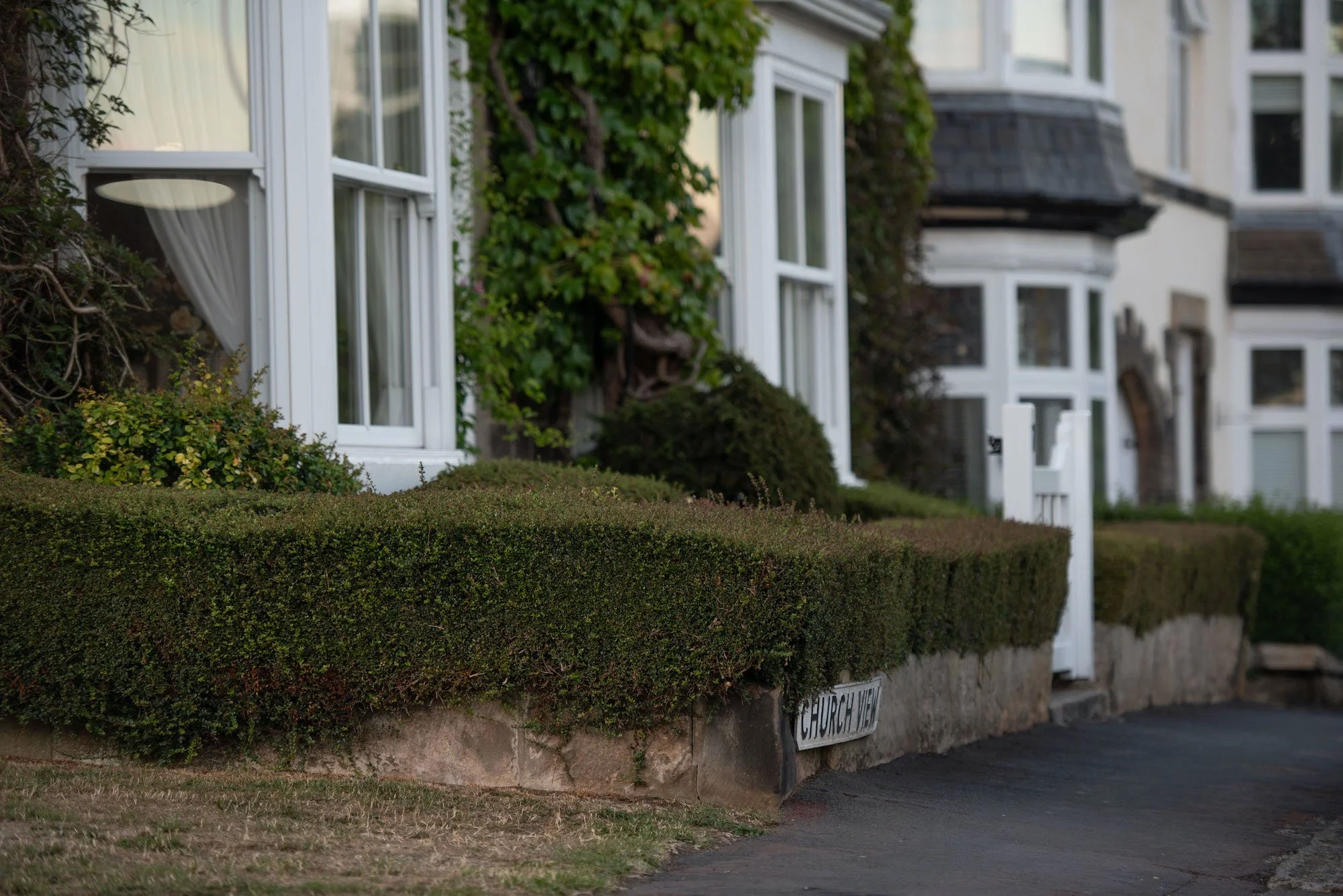 Row of white houses with bay windows, surrounded by neatly trimmed bushes and ivy climbing on the walls. A street sign reads 'Church View' in front of the buildings.
