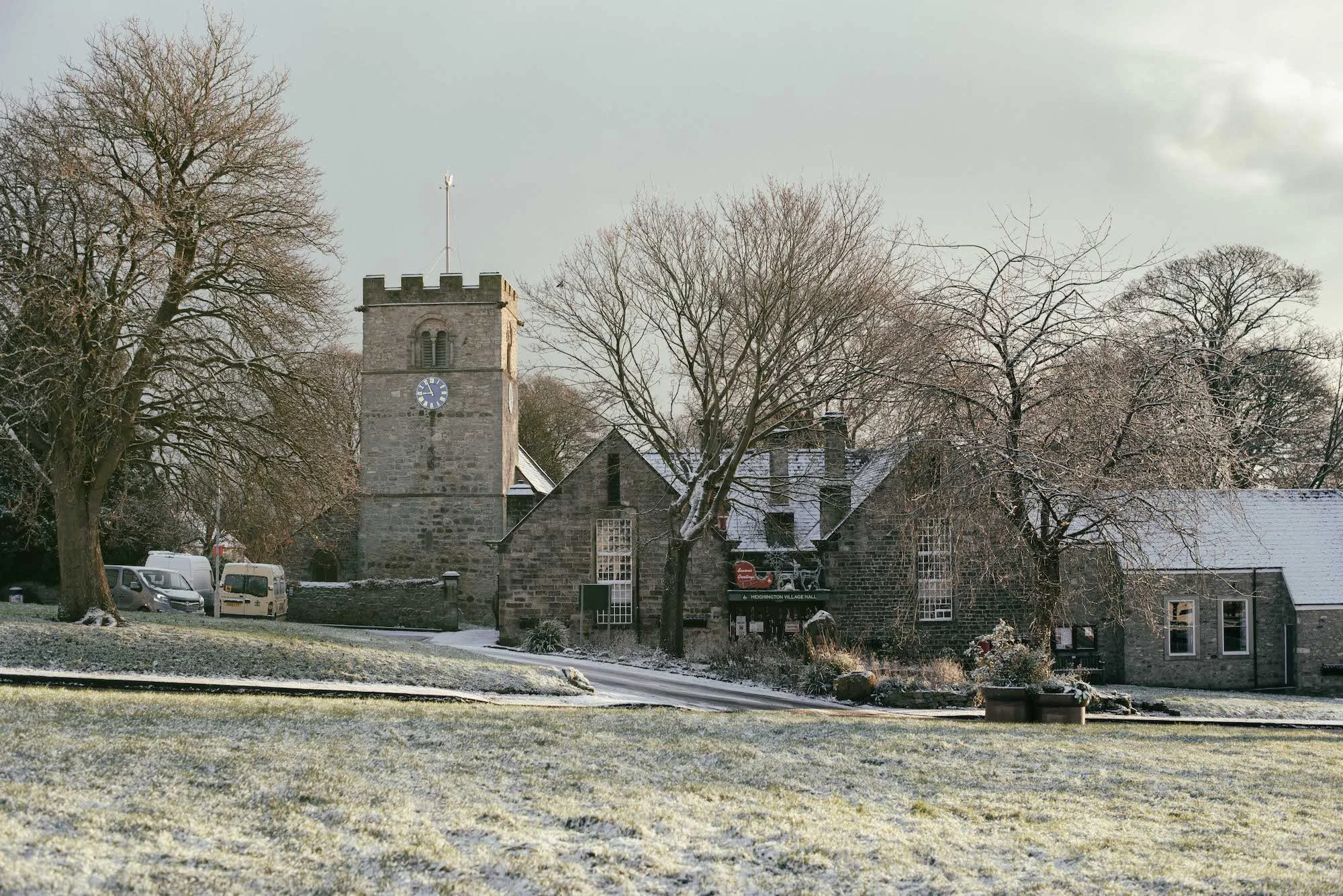 St. Michaels church in Heighington at winter time. Snow-covered grass in the foreground with stone church and bare trees behind, overcast sky in the background.