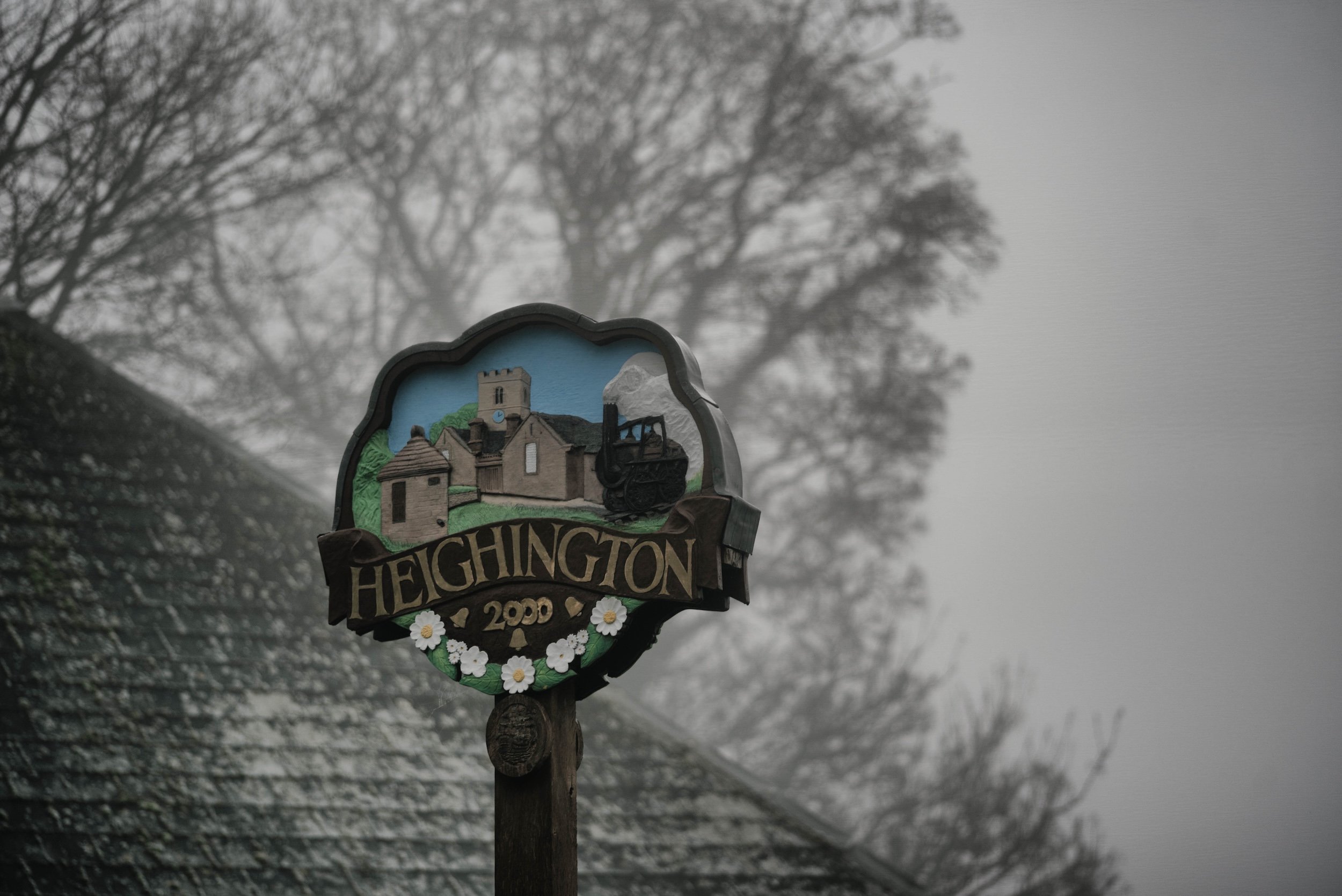 A decorative signpost reading 'HIGHTINGTON 2020', featuring a painted background of a church and a landscape, attached to a wooden post. In the background, there are leafless trees and a cloudy sky, indicating a winter or fall setting.