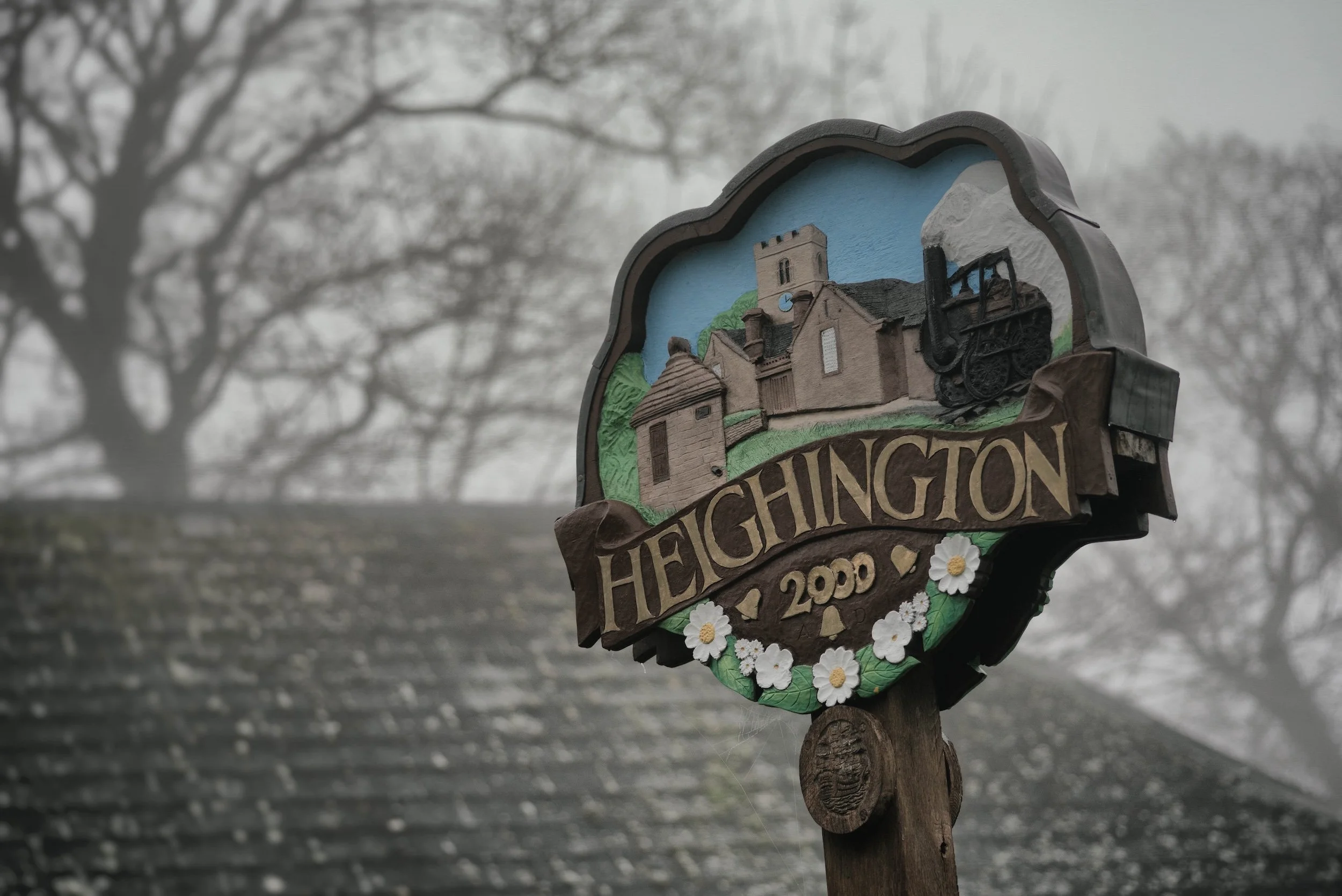 A decorative signpost shows the town of Hechingen, with a painted scene of a church and landscape, and the year 2000 at the bottom, surrounded by small white flowers. The background is foggy with leafless trees.