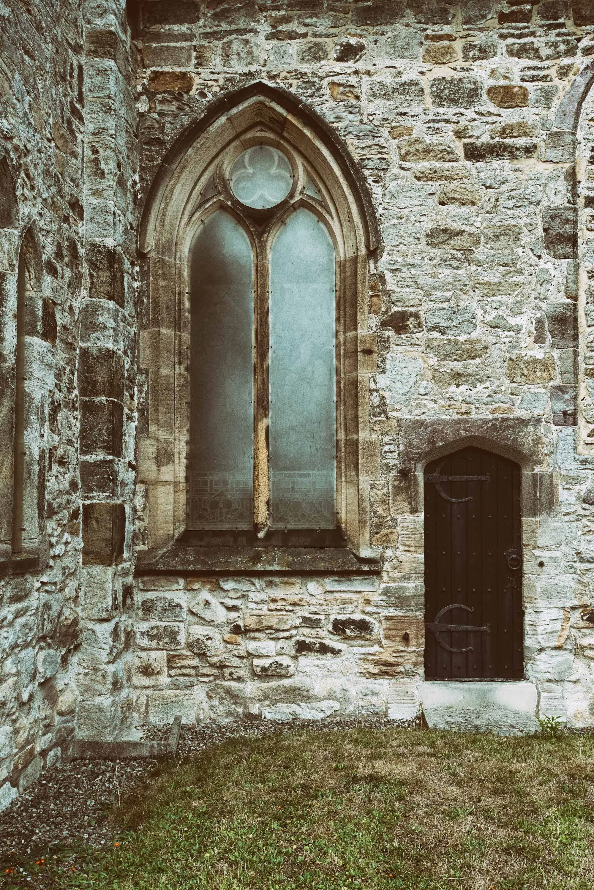St. Michael's Church wall with stained glass window and small wooden door, surrounded by grass and plants.