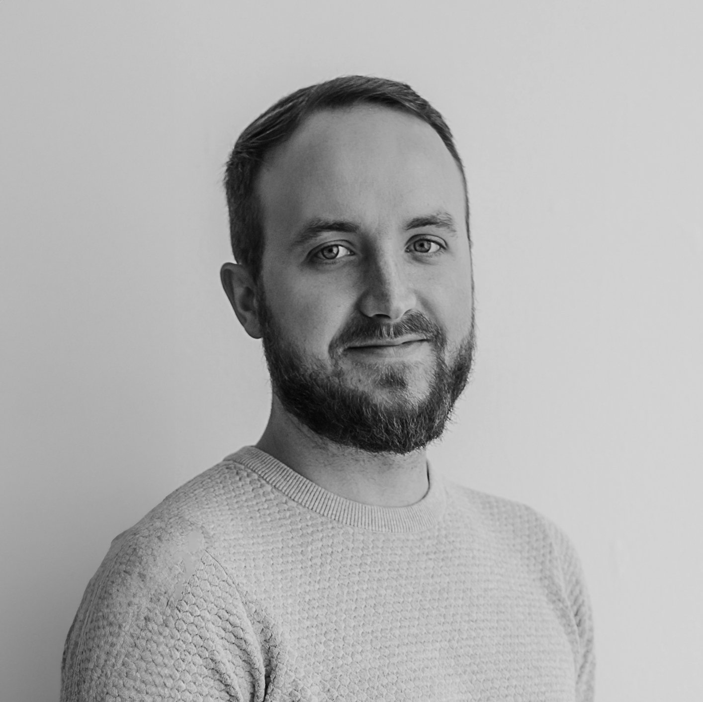 Black and white portrait of a man with a beard and short hair, wearing a textured sweater, looking at the camera with a slight smile, against a plain light background.