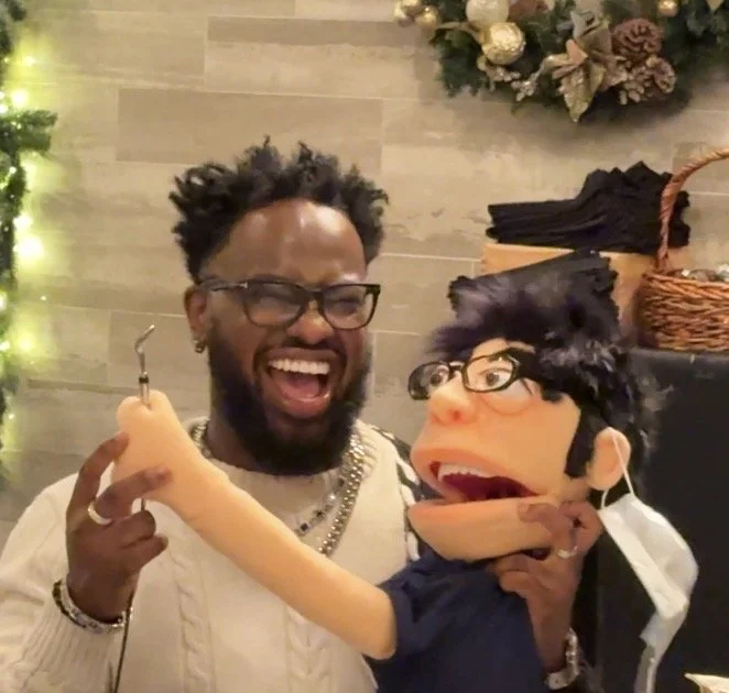 A man laughing and holding a puppet with glasses and black hair, in a Christmas-themed setting.