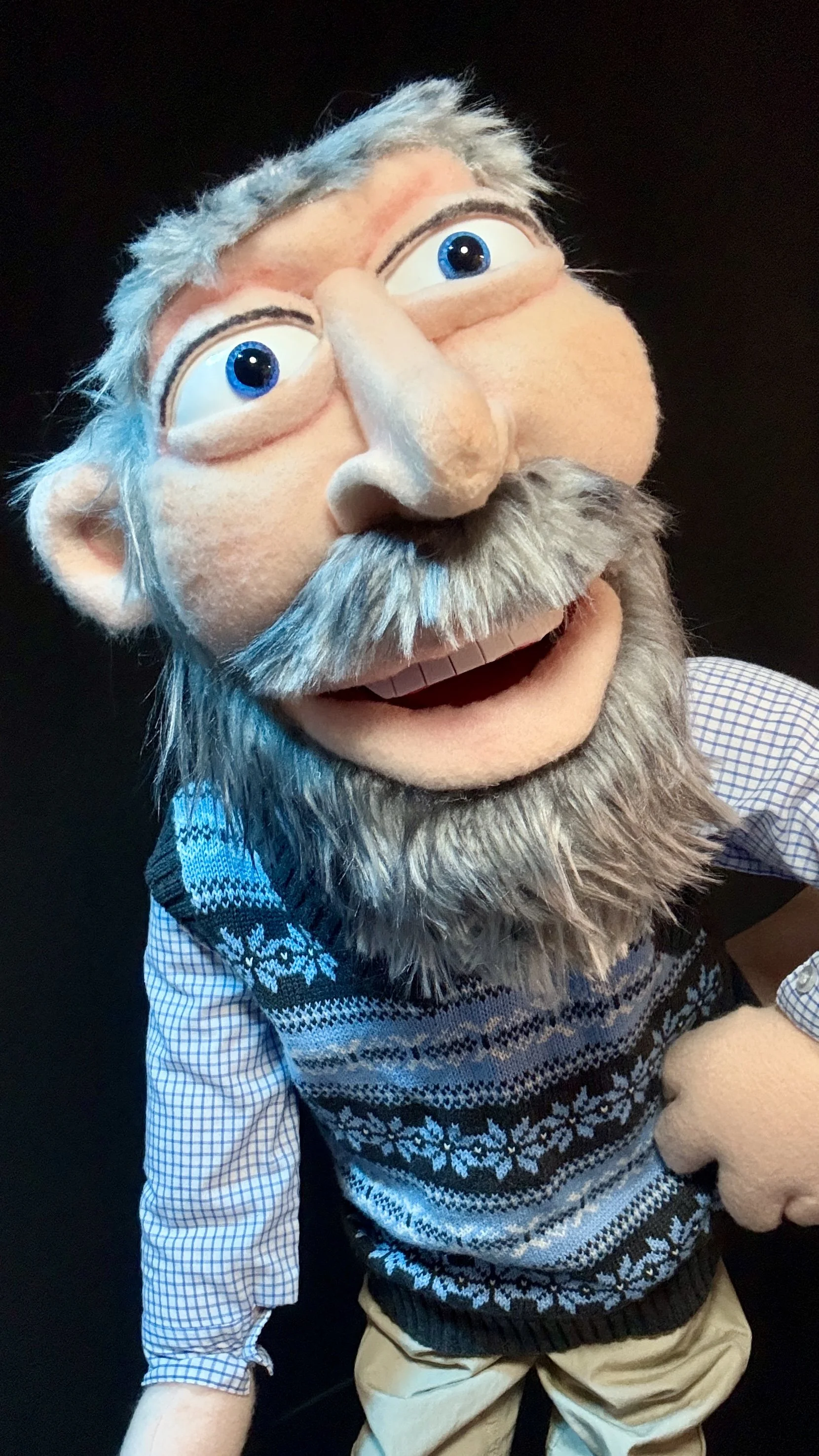 Close-up of a puppet with gray hair, blue eyes, a large nose, a beard, and mustache, wearing a blue and white patterned sweater and beige pants, against a black background.