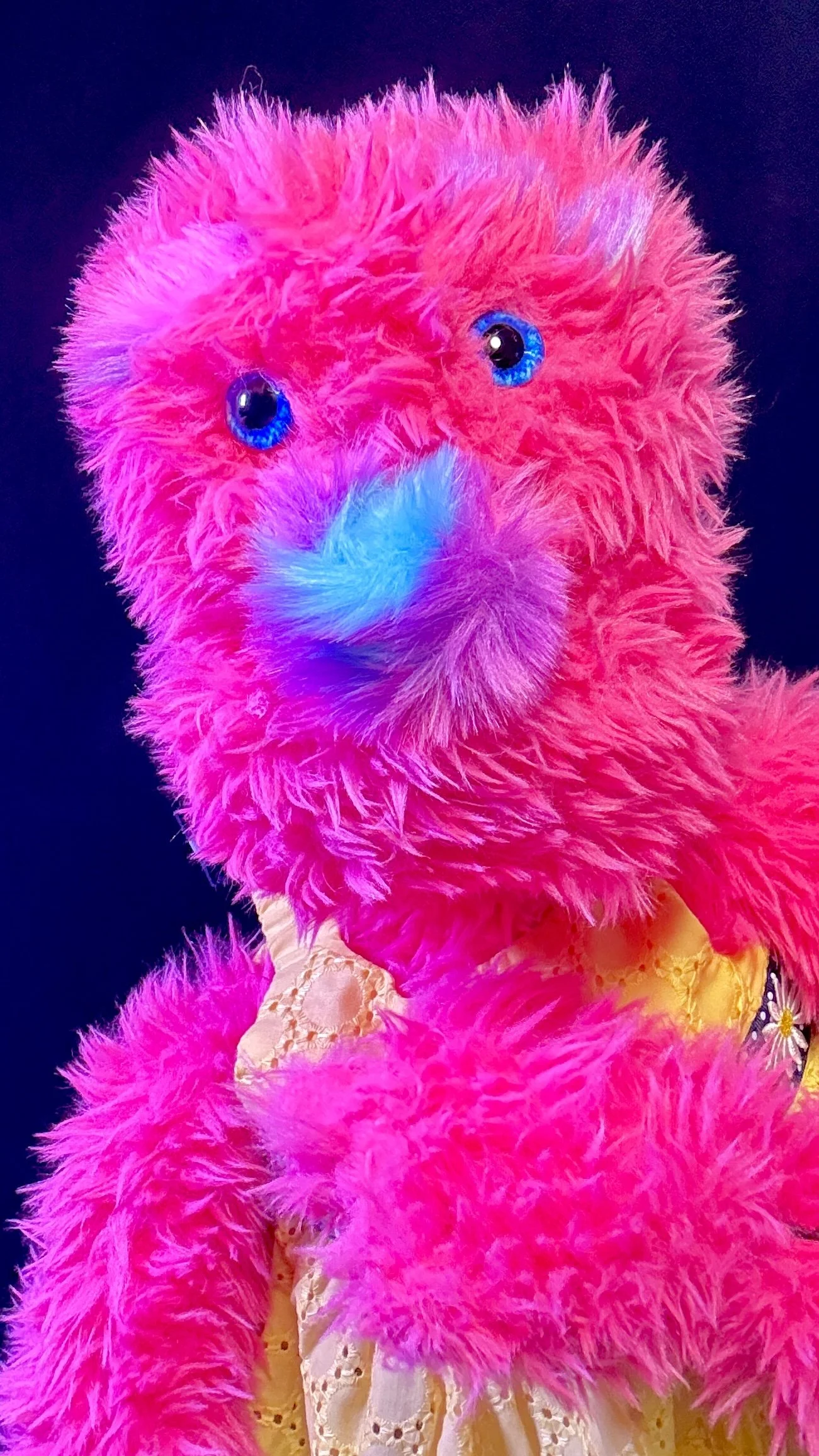 Pink furry puppet with blue eyes and a glowing blue nose, wearing a yellow dress with eyelets.