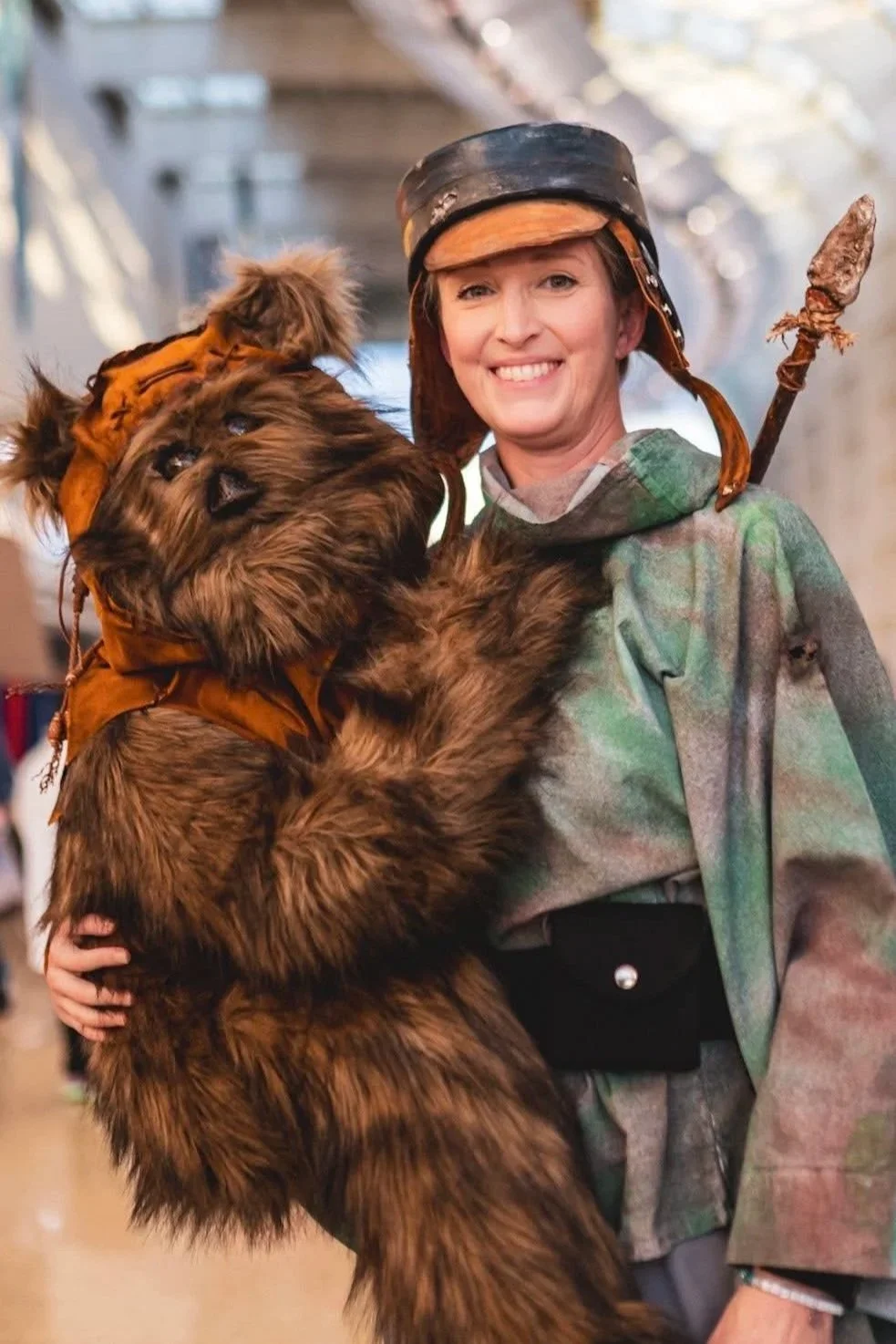 Woman dressed as a hunter in a camouflage jacket, wearing a helmet, holding a large teddy bear dressed as a hunter with a bushy fur coat and a small hat, with a background of an indoor setting.