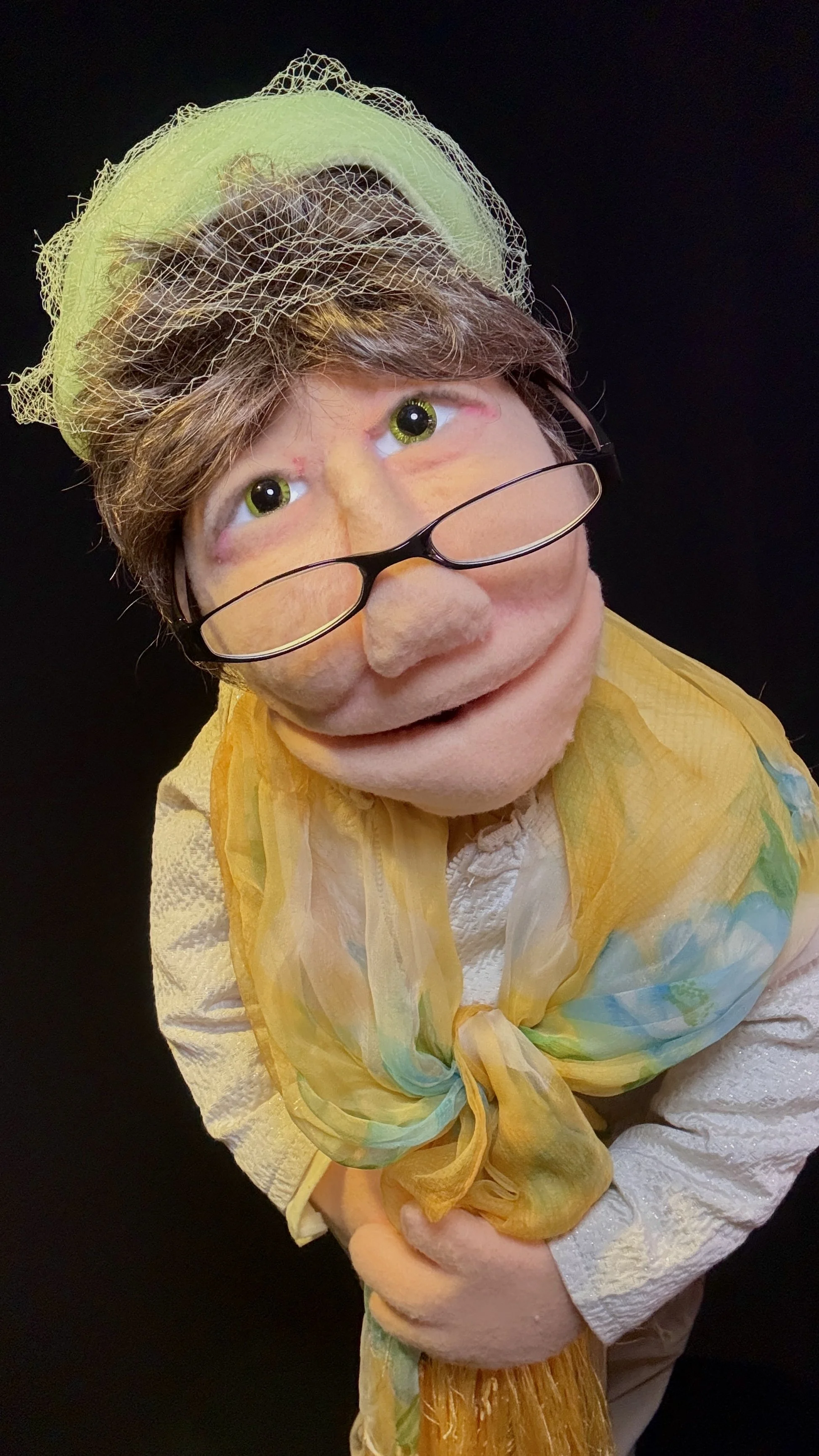 A puppet of an elderly woman with glasses, green eyes, a hat with netting, a yellow scarf, and a white blouse, smiling against a black background.