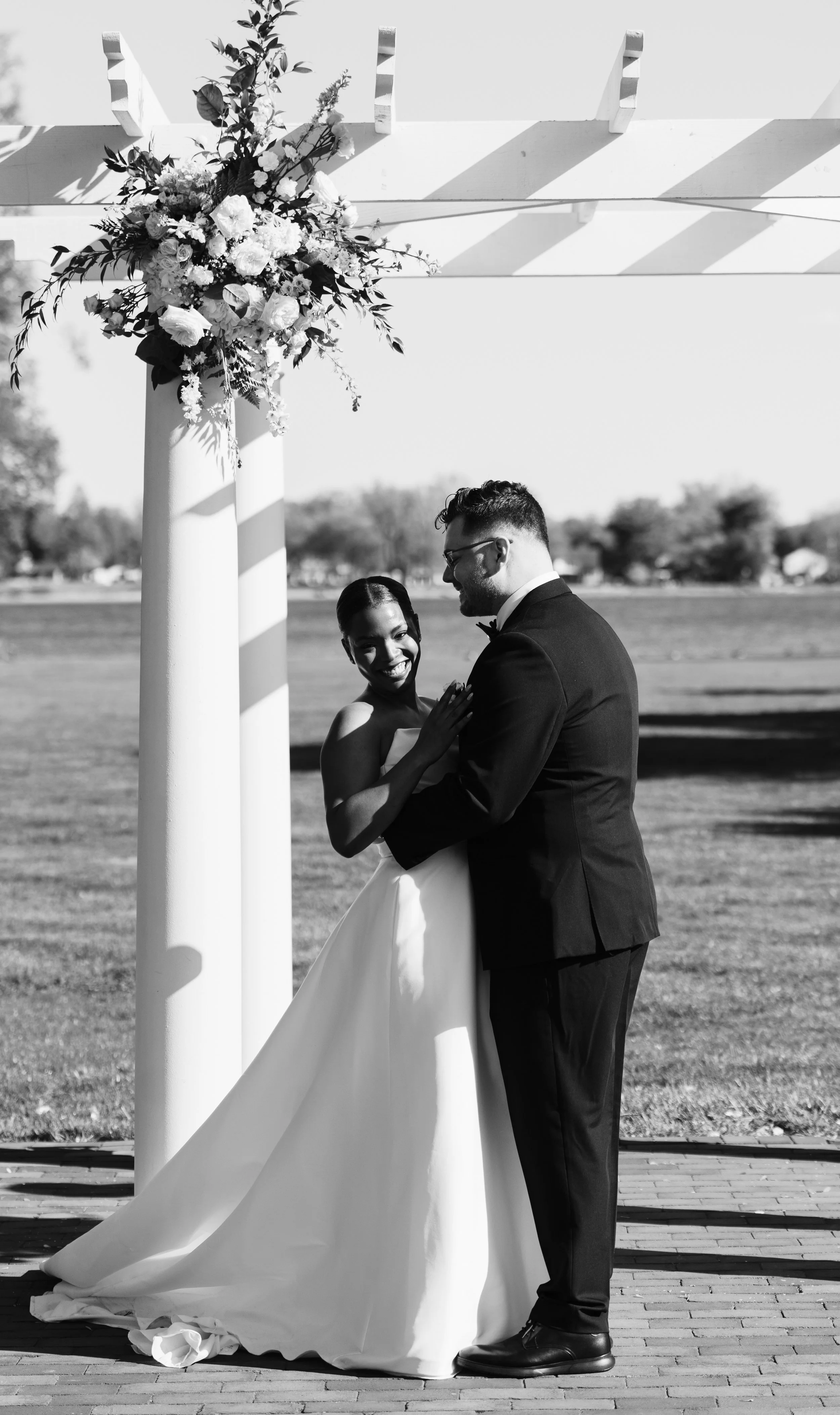A bride and groom share a dance under a decorated wedding arch outdoors, with the bride smiling and the groom in a tuxedo, in black and white photograph.