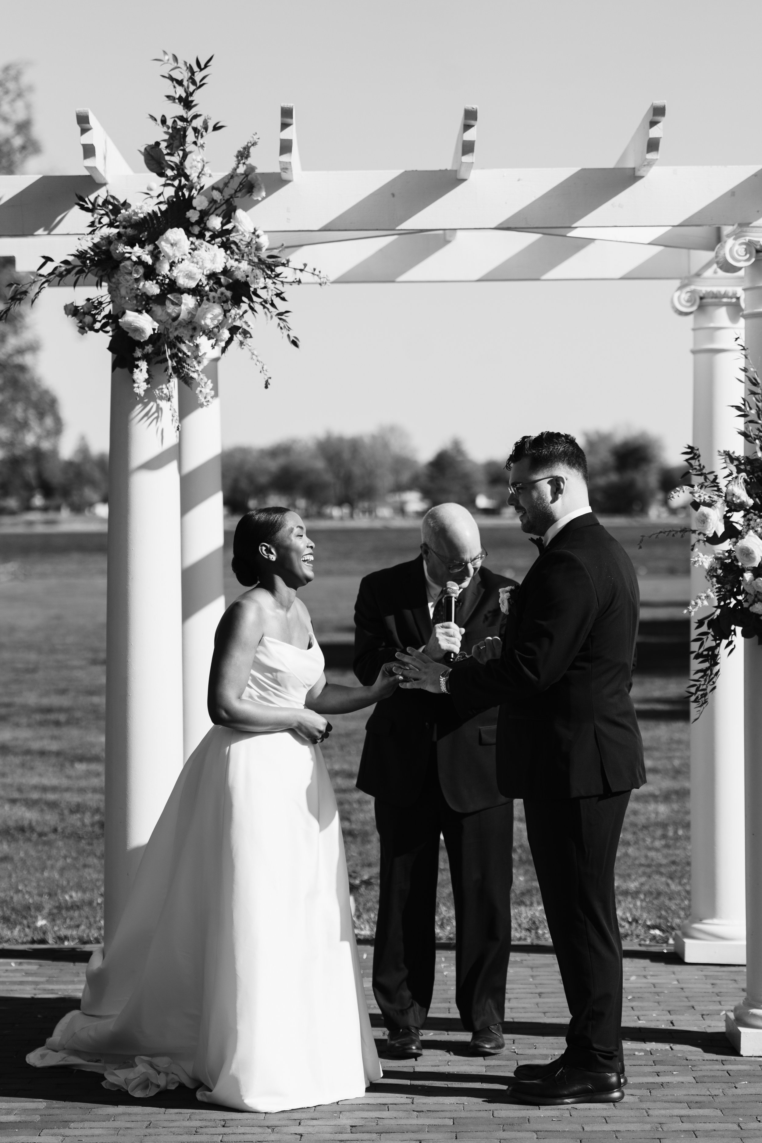 A wedding ceremony outdoors under a decorated arbor with a bride and groom exchanging rings, officiant holding a microphone, on a sunny day.