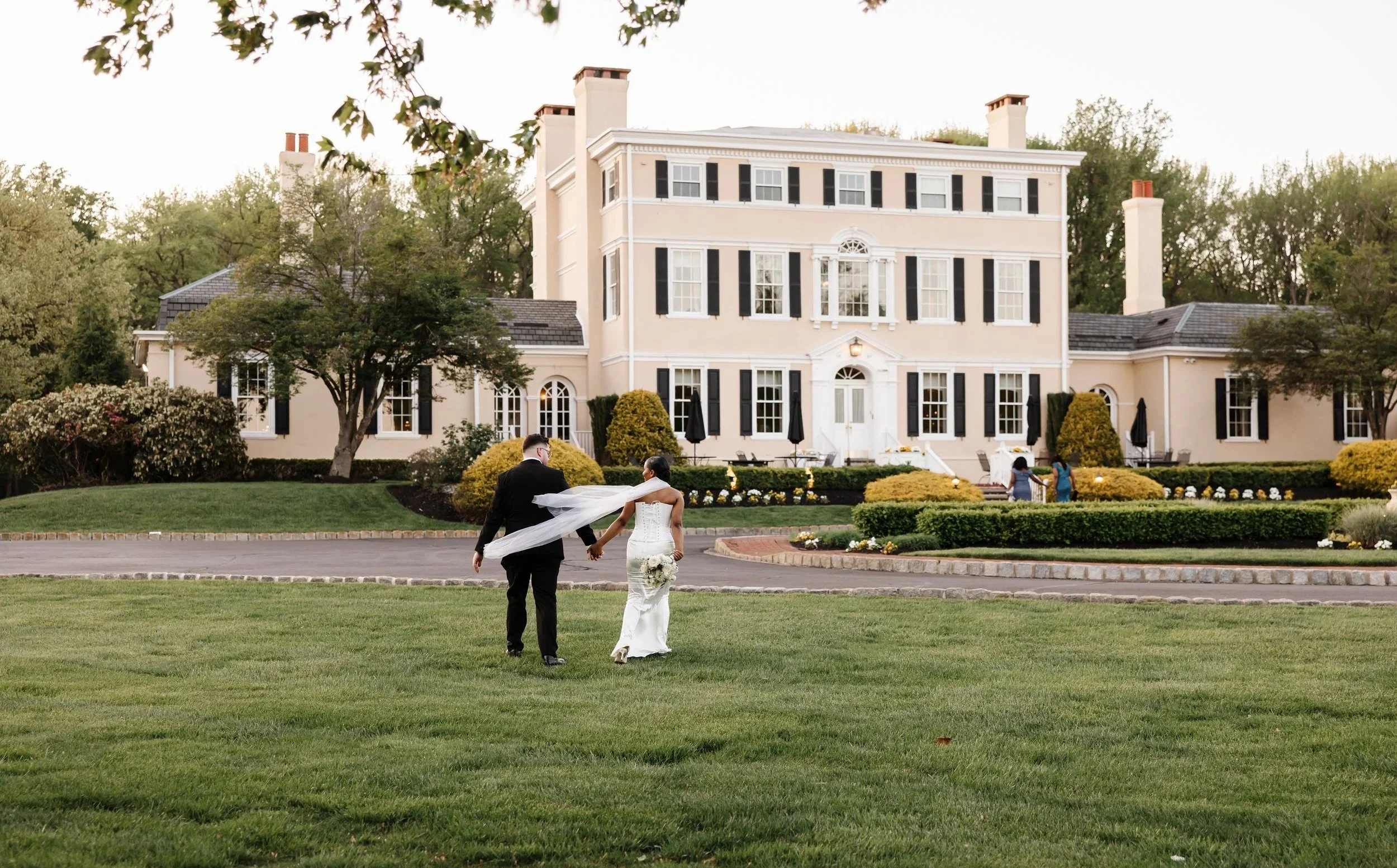 A newlywed couple holding hands and walking on a lawn in front of a large, elegant, cream-colored mansion with black shutters, surrounded by trees and landscaped greenery.
