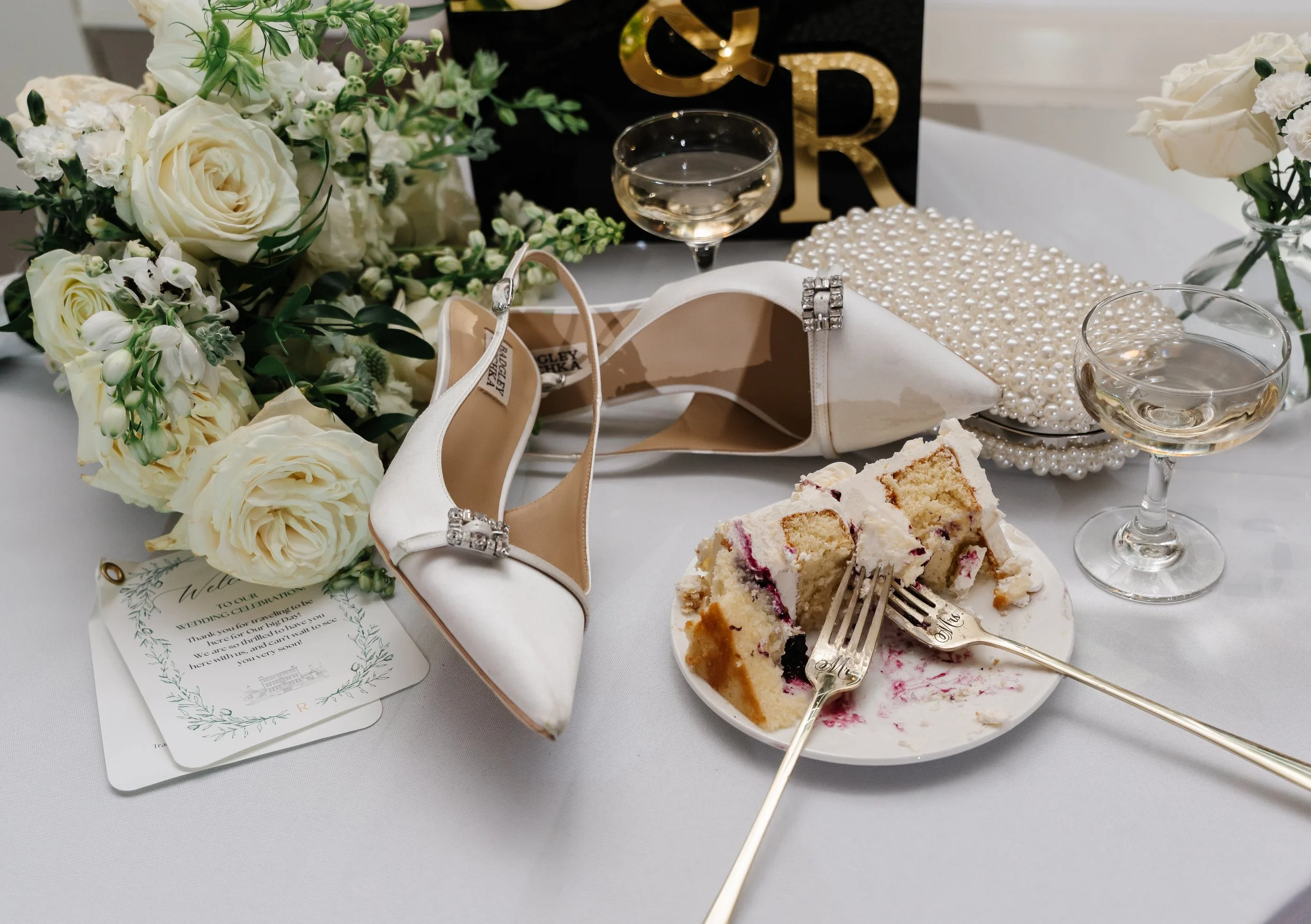 A wedding reception table with a floral centerpiece of white roses and greenery, a pair of white high-heeled shoes with embellishments, a pearl-embellished clutch, two glasses of white wine, a partially eaten piece of wedding cake with a fork and knife, and wedding cards.