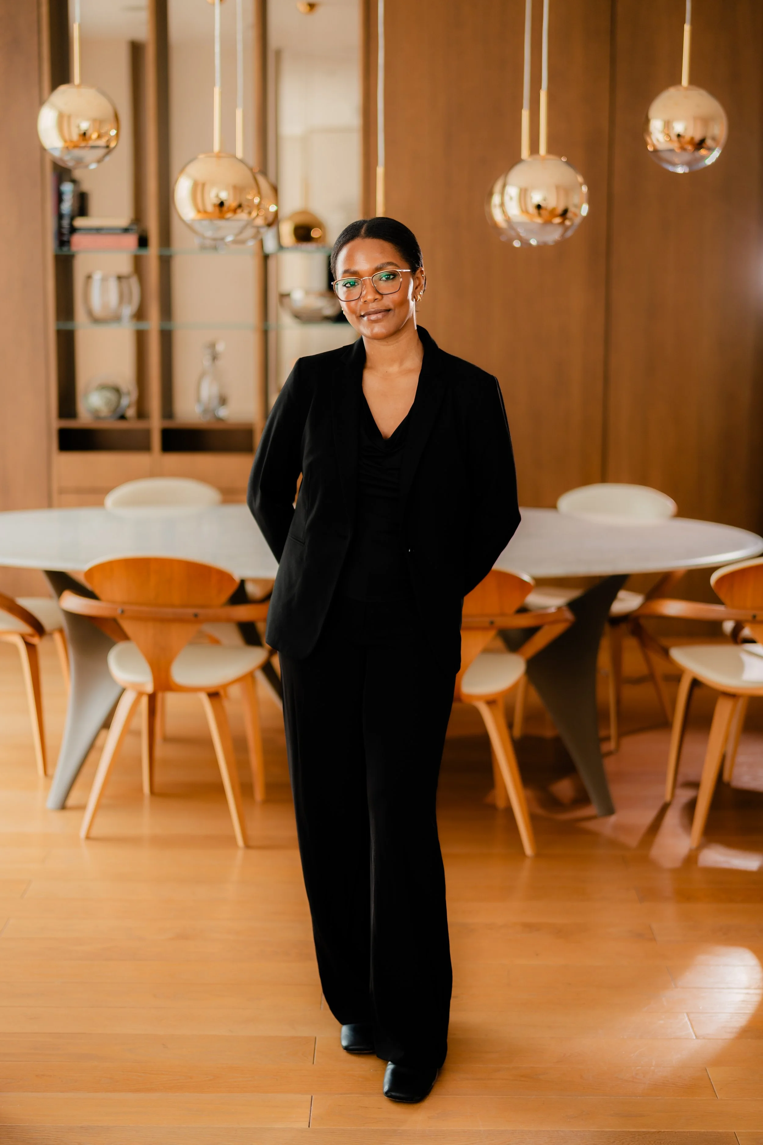 A woman dressed in a black suit standing in a modern, well-lit conference room with wooden walls and light-colored chairs around a round table.