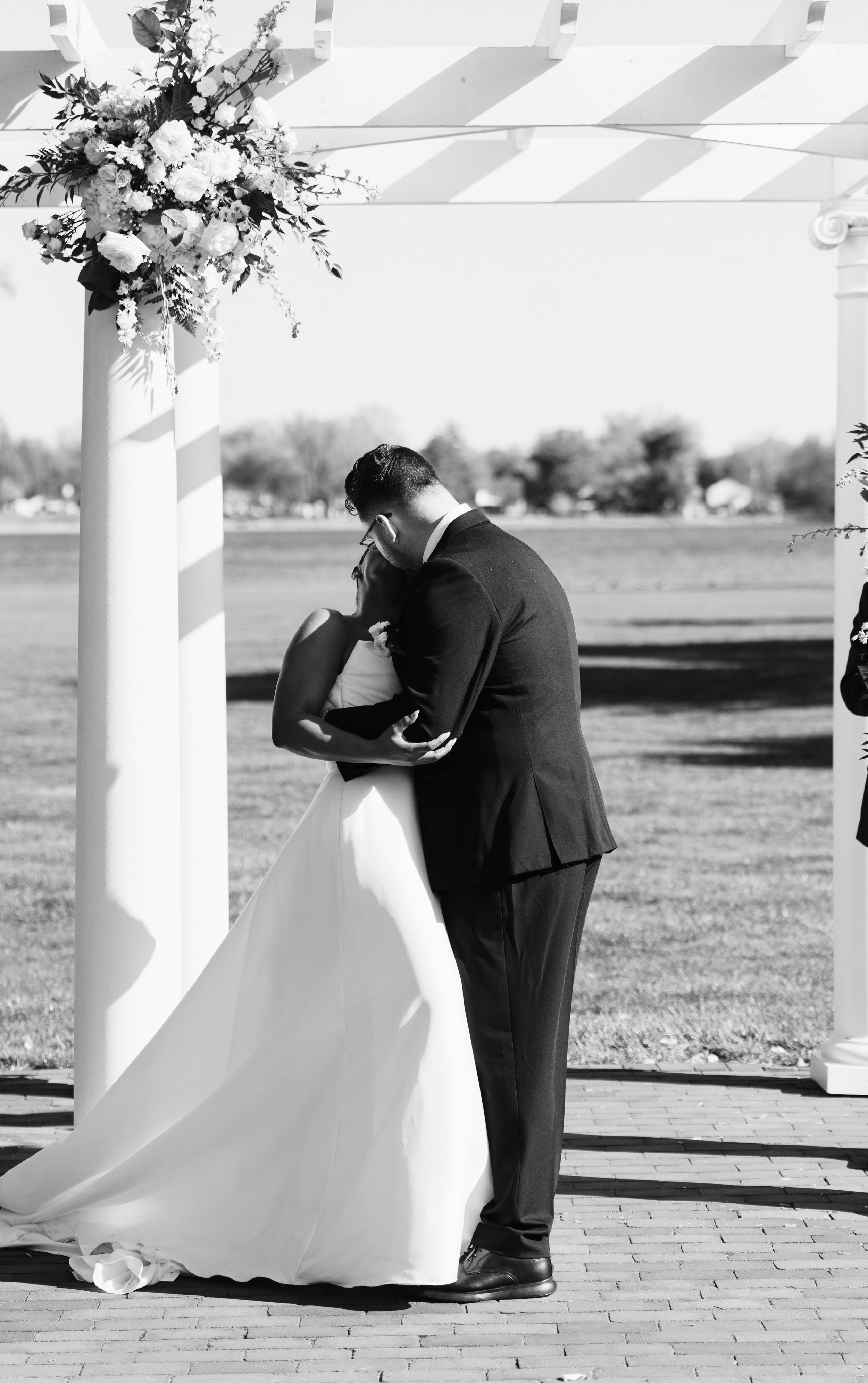 A black-and-white photo of a bride and groom sharing a kiss under a wedding arch with floral decorations in an outdoor setting.