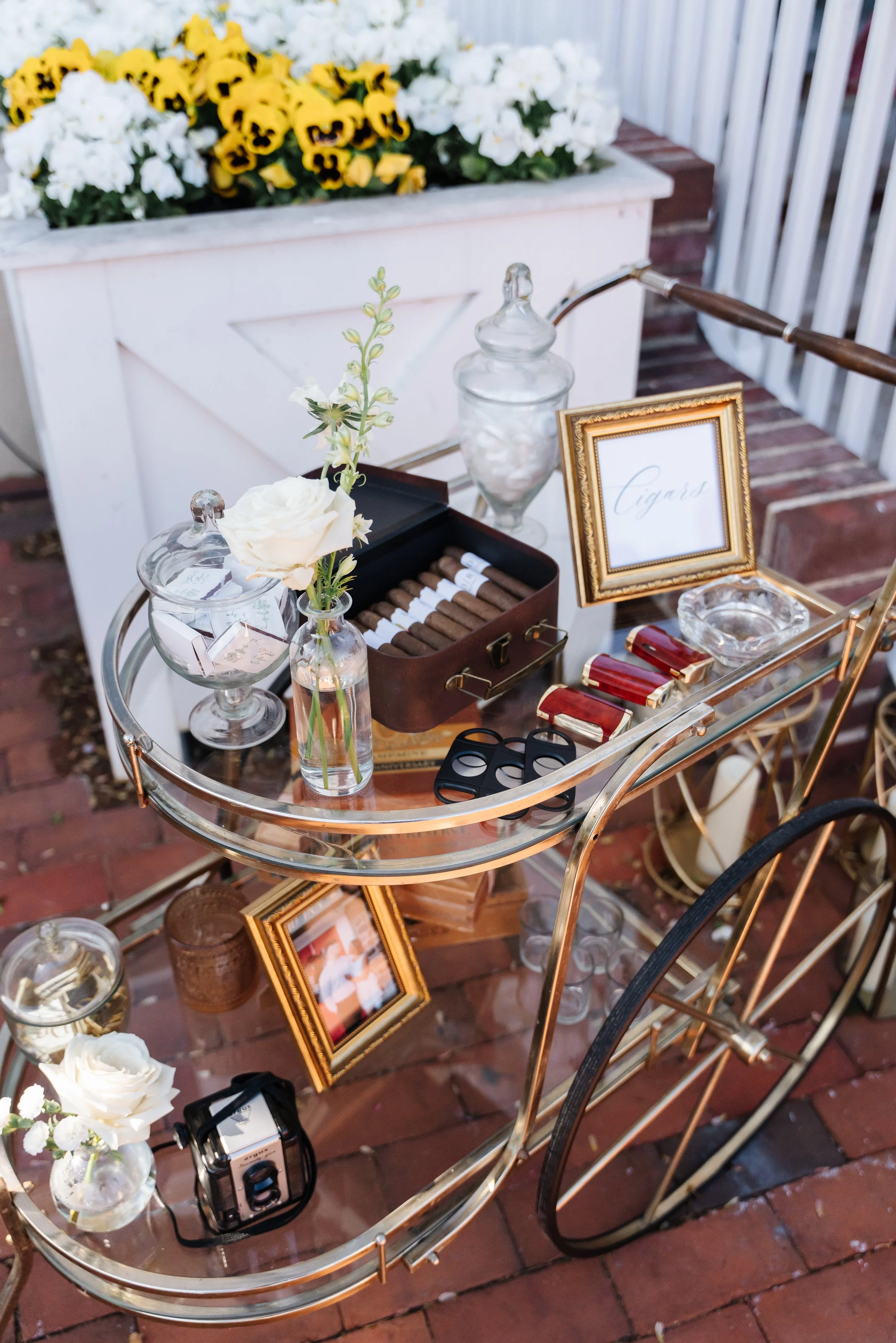 A vintage gold bar cart with cigars and accessories, decorated with white roses and flowers in glass vases, set on a brick patio with a flower box filled with white and yellow pansies in the background.