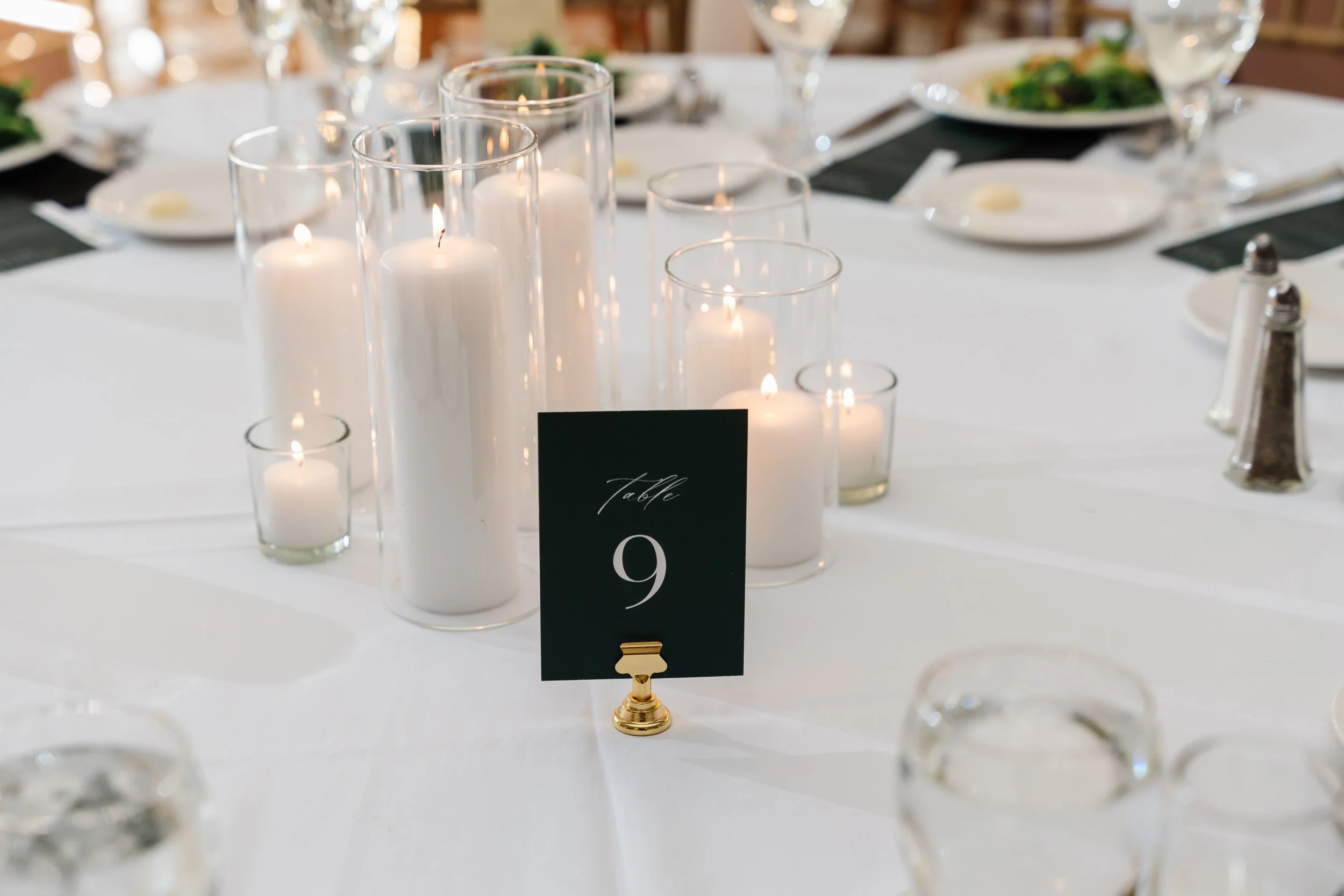 Table decorated with white candles in glass candle holders and a black table number card with gold clip reading 'Table 9' at a formal event.