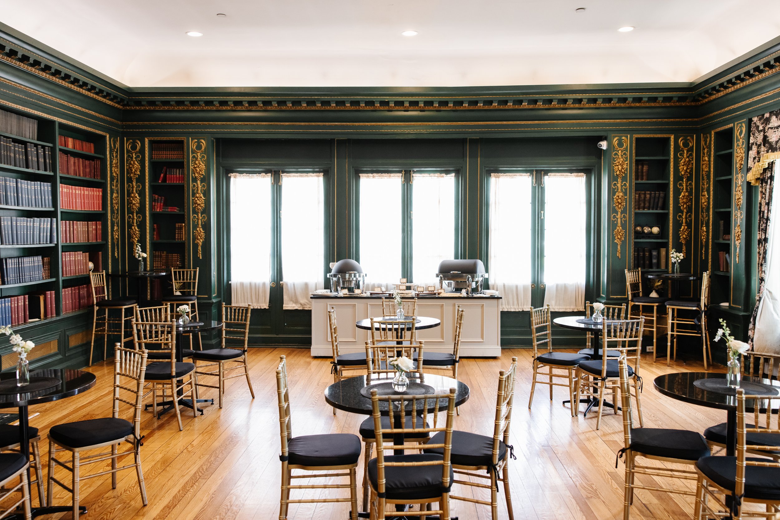 Elegant room with dark green walls adorned with gold accents, tall windows with white curtains, wooden floor, round tables with black tops, gold chairs with black cushions, and a buffet table at the back.
