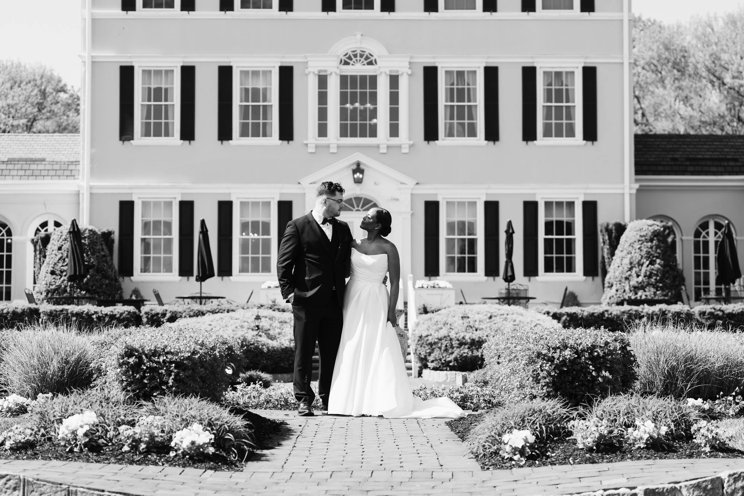 A couple, possibly newlyweds, standing on a garden pathway in front of a large, elegant house with multiple windows and shutters. The woman is wearing a white wedding dress and holding a small bouquet, while the man is in a dark suit. The scene appears to be a wedding or engagement photo.