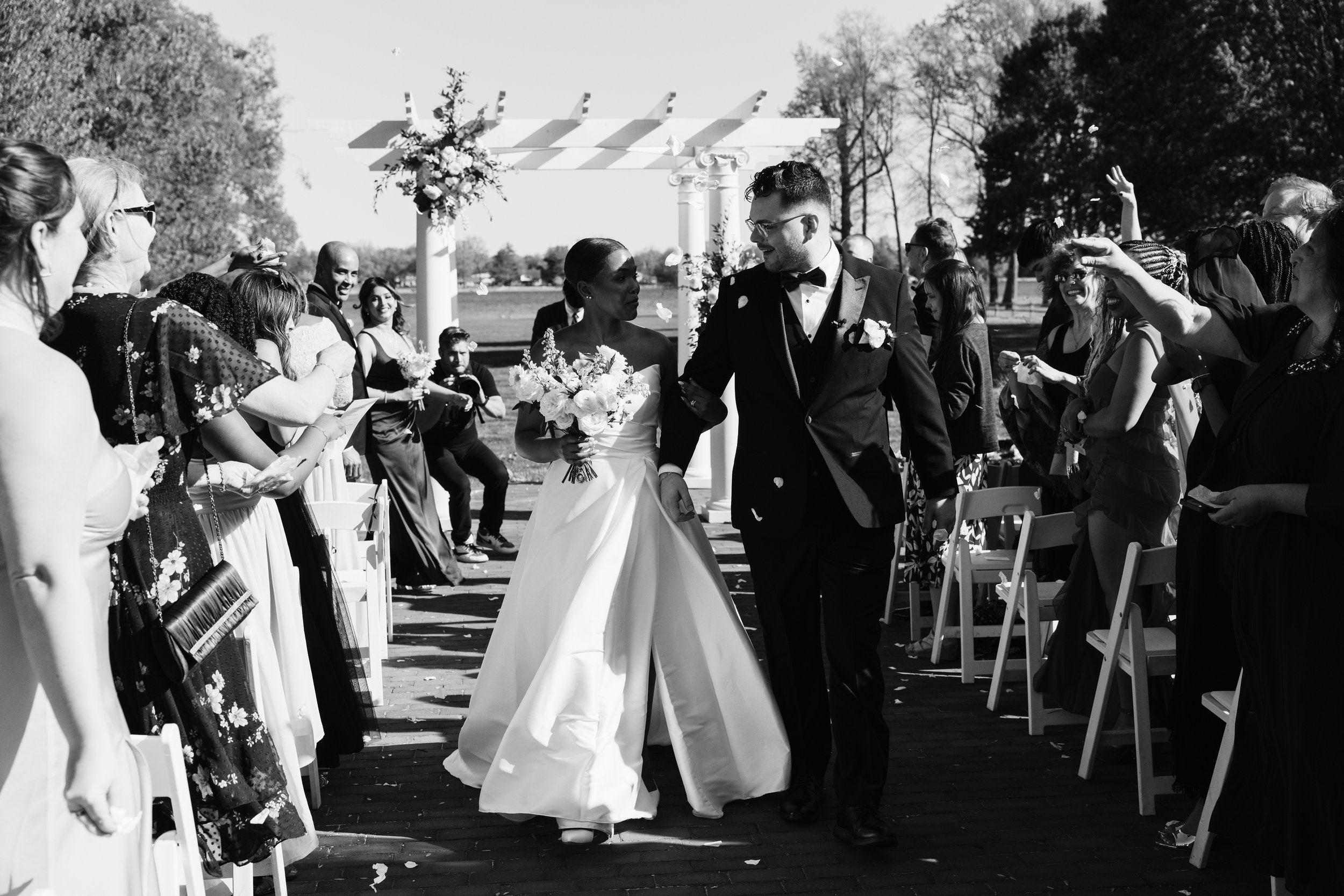 Black and white photo of a newlywed couple walking down an outdoor aisle, holding hands. The bride is in a white wedding dress holding a bouquet, and the groom in a tuxedo. Guests are on both sides, smiling, clapping, and throwing flower petals. Ther