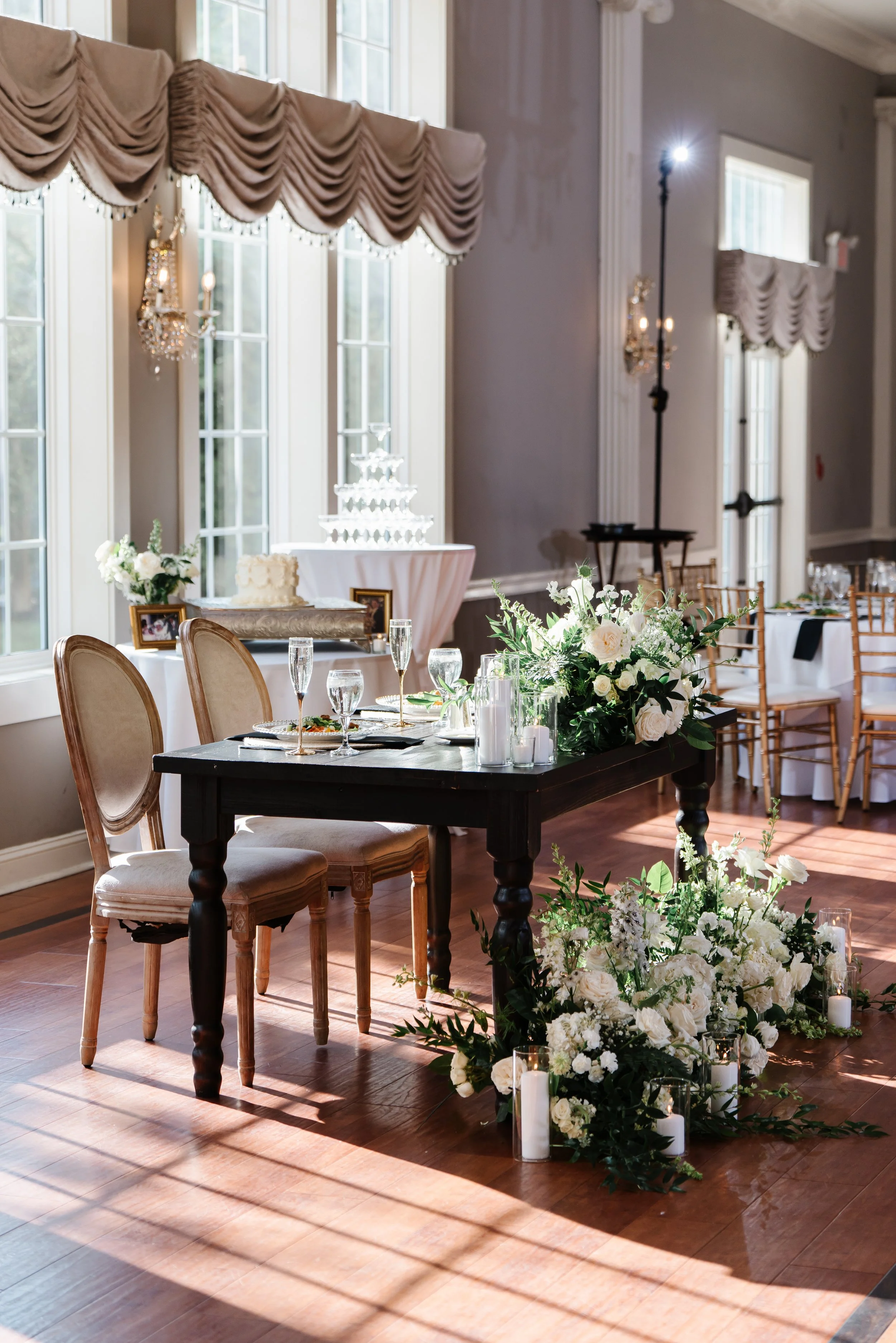 Elegant indoor wedding reception setup featuring a long black table decorated with white floral arrangements and candles, surrounded by beige upholstered chairs, with a decorated cake table and champagne on the side, large windows with draped curtains, and natural sunlight.