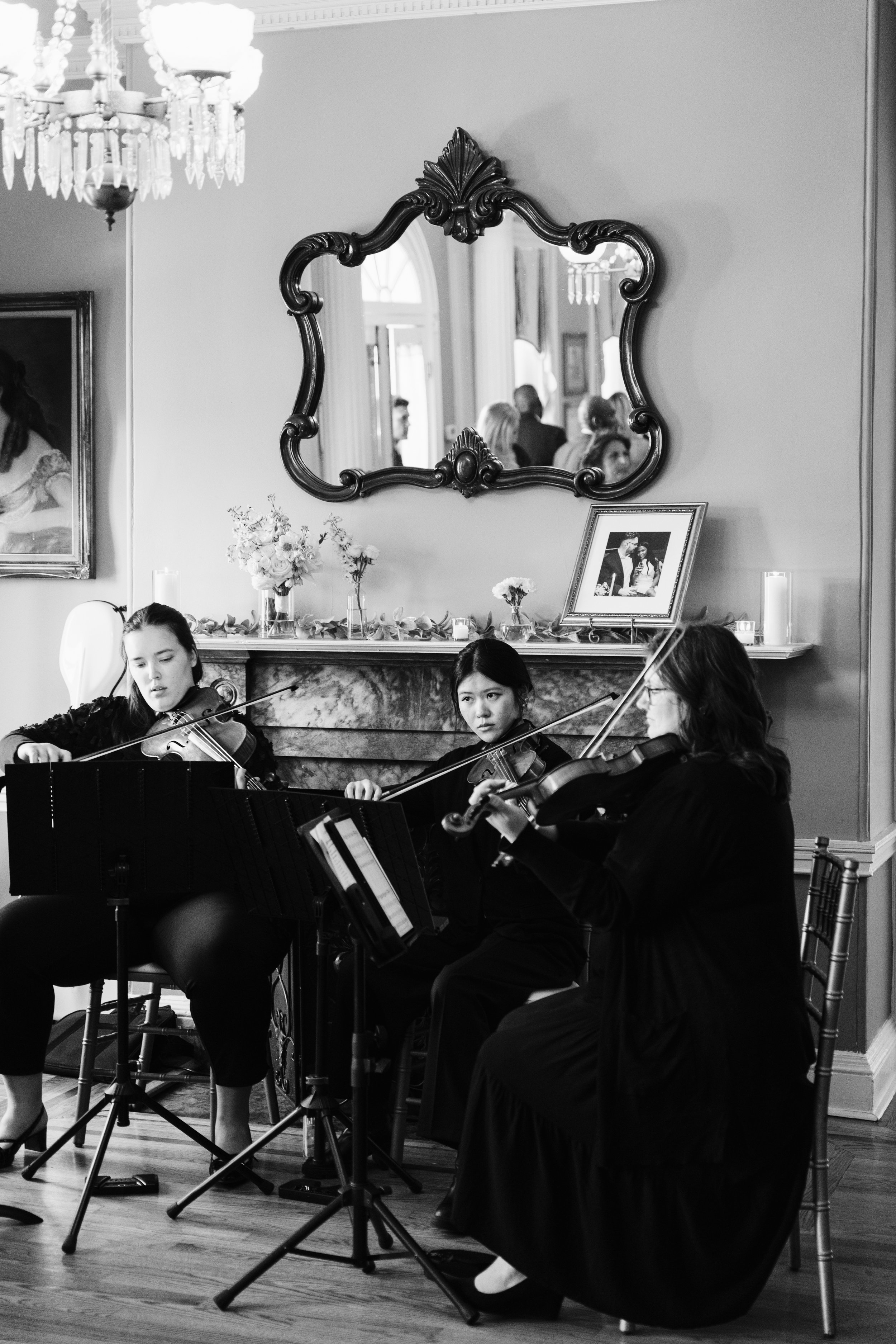 Three women playing violins and a viola in an indoor setting, with a mirror, framed photo, and decorative items on a mantle behind them, and an audience reflected in the mirror.