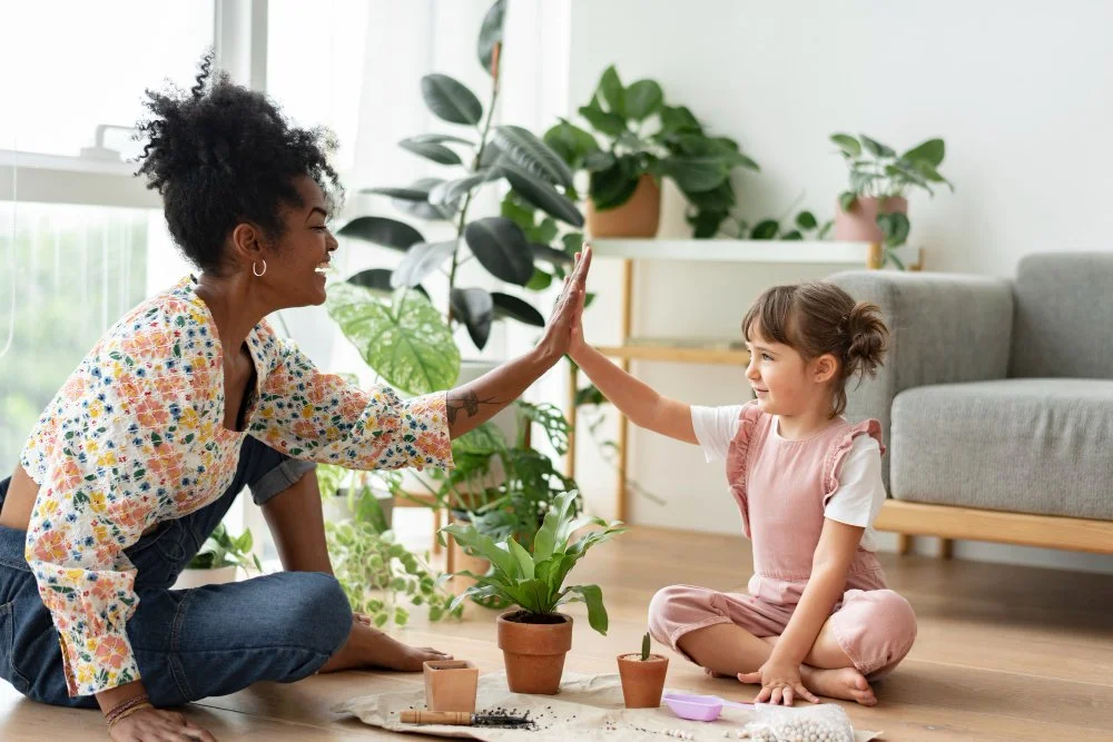 A woman and a young girl sitting cross-legged on the floor inside a living room, giving each other a high five. There are houseplants around them and a gray couch in the background.