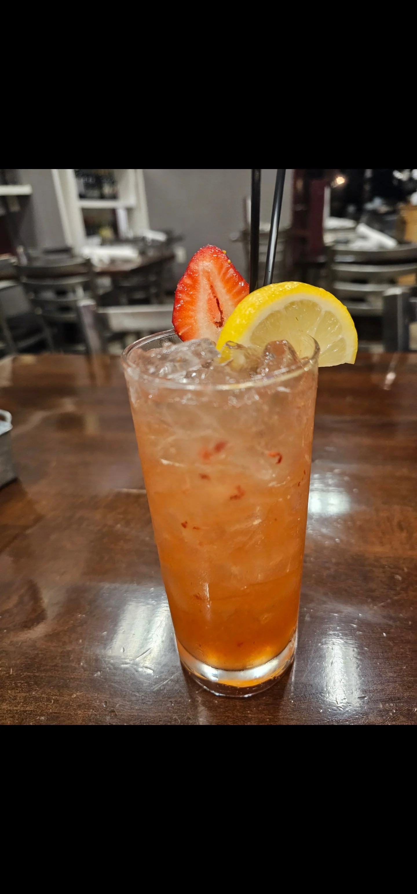 A tall glass of iced strawberry lemonade garnished with a sliced strawberry and a lemon wedge, with black straws inside, on a wooden table in a restaurant setting.