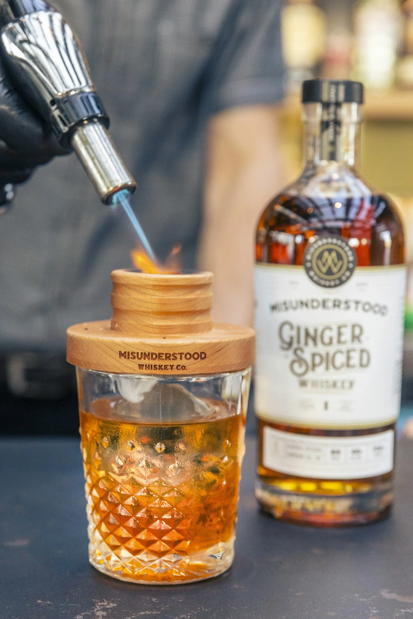 A person using a culinary torch to caramelize sugar in a glass with ice and whiskey, with a bottle of Misunderstood Ginger Spiced Whiskey in the background.