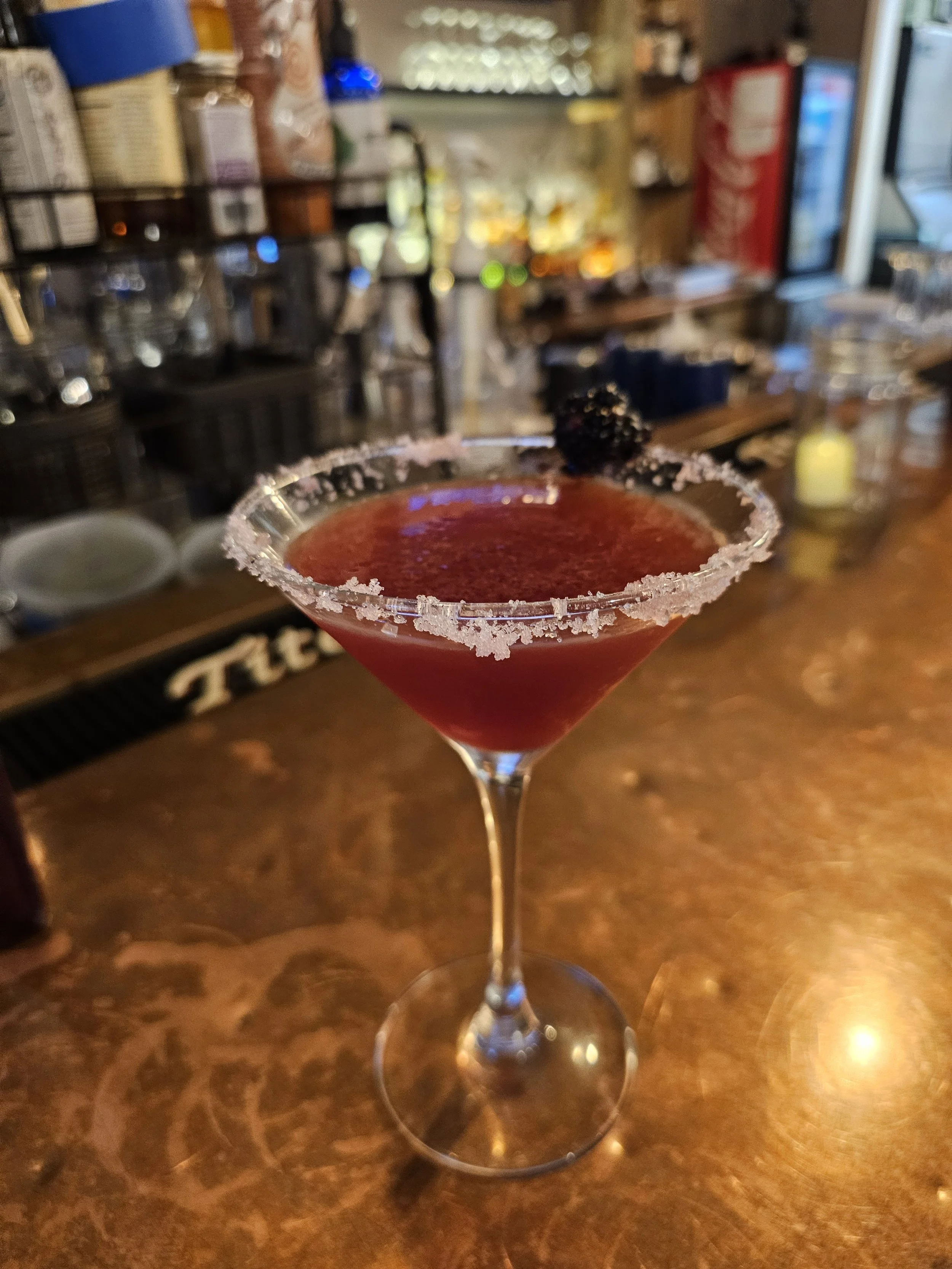 A red cocktail in a martini glass with a salted rim and a blackberry garnish on top, set on a bar counter.