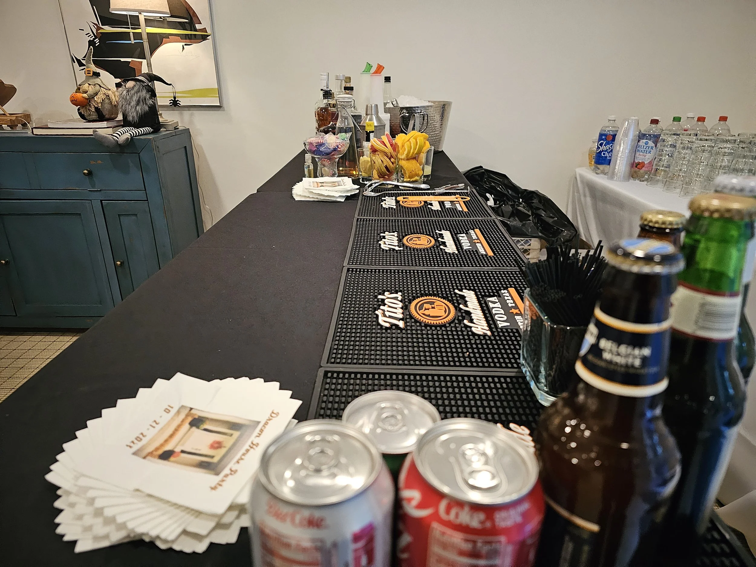 Bar counter with drink cans, beer bottles, and napkins, with a bar mat displaying the logo for 'the lot' and a table of bottled beverages in the background. There are chocolate and caramel candies and various drinks, including water bottles, on the s