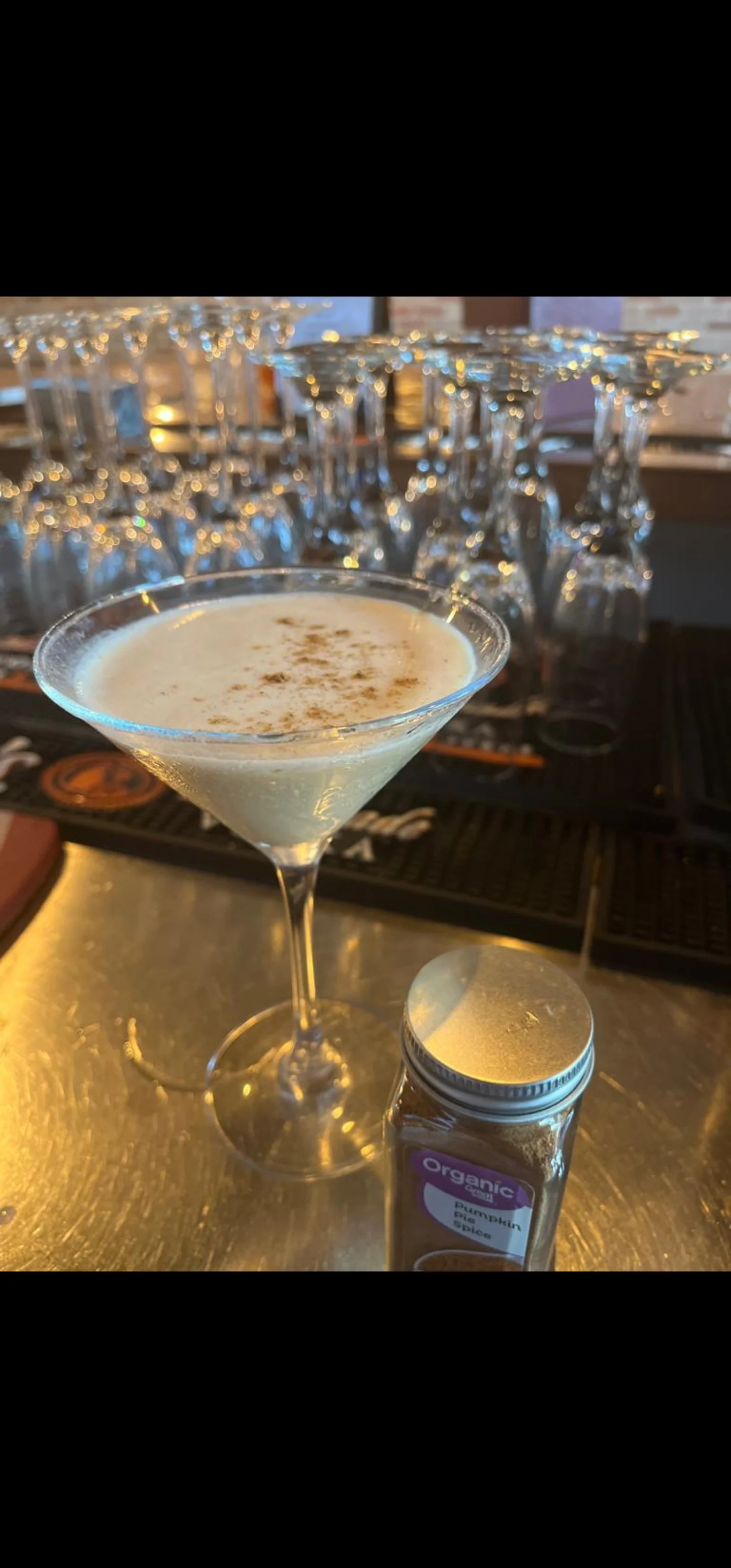 A martini glass filled with a creamy cocktail topped with spices, placed on a bar counter with a jar of pumpkin spice next to it, and a background of upside-down glasses.