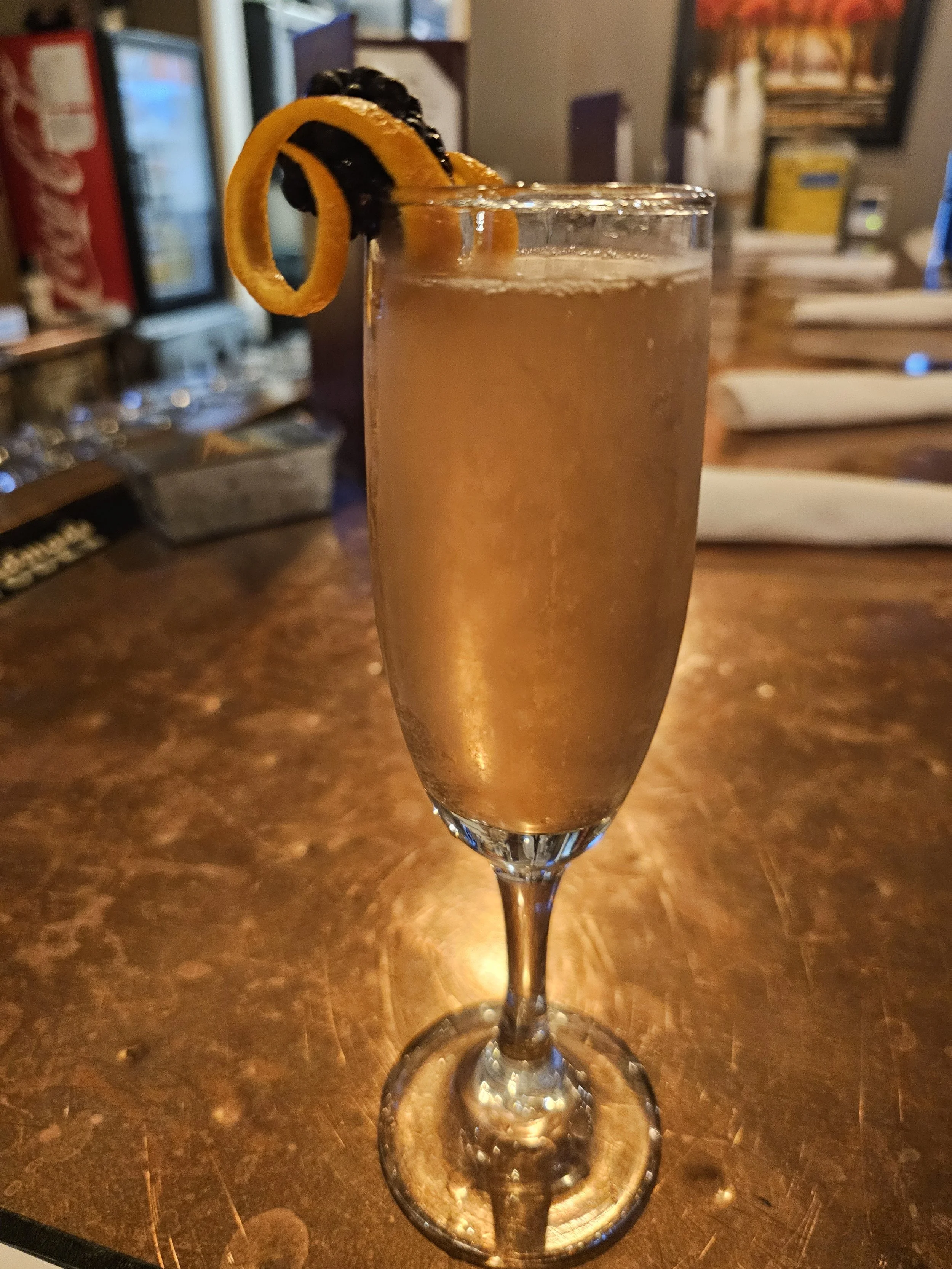 A tall champagne flute filled with a sparkling, golden-colored cocktail garnished with orange twist and black cherry peel.