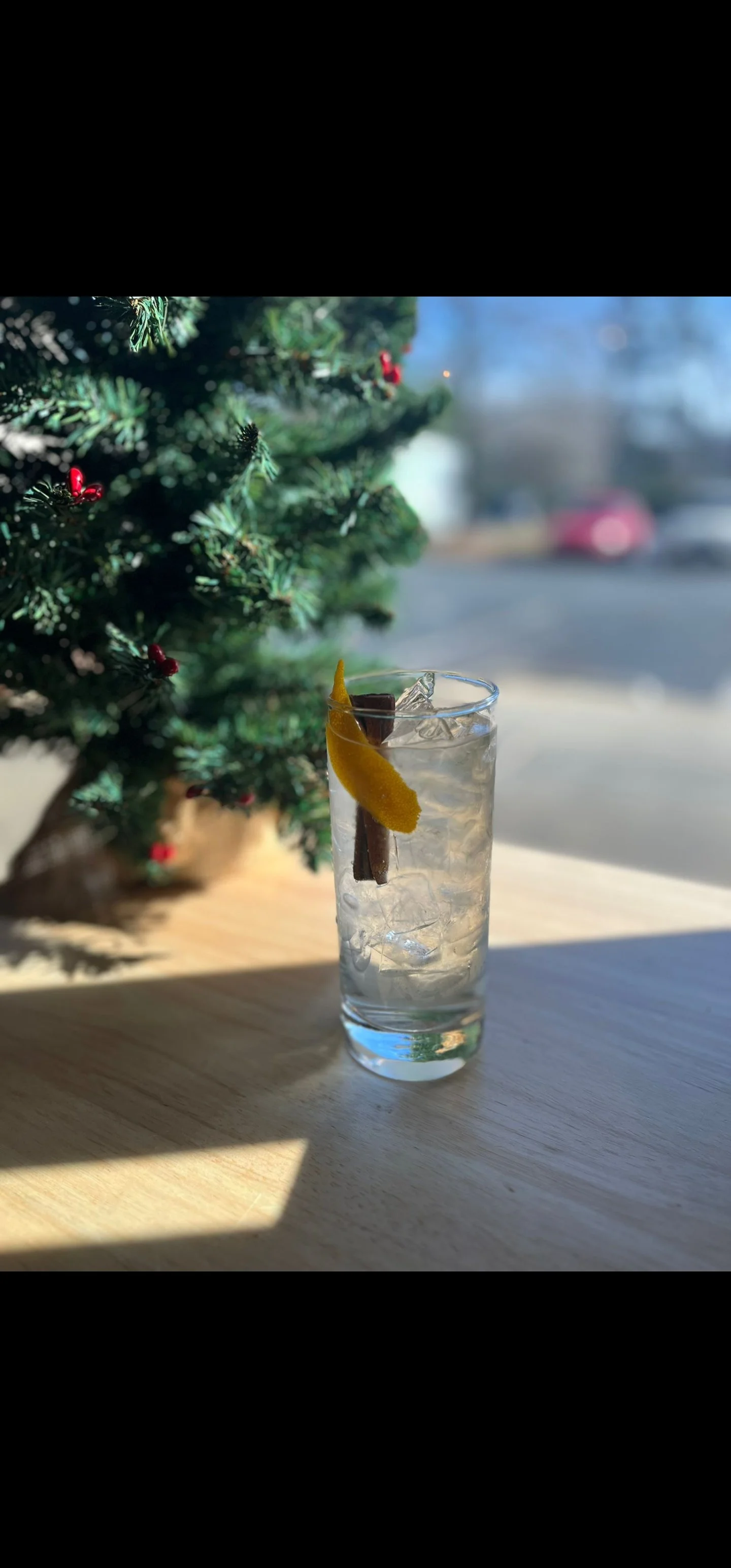 A tall glass of what appears to be a cocktail with ice, garnished with a lemon peel and a cinnamon stick, placed on a wooden surface near a decorated Christmas tree outside.