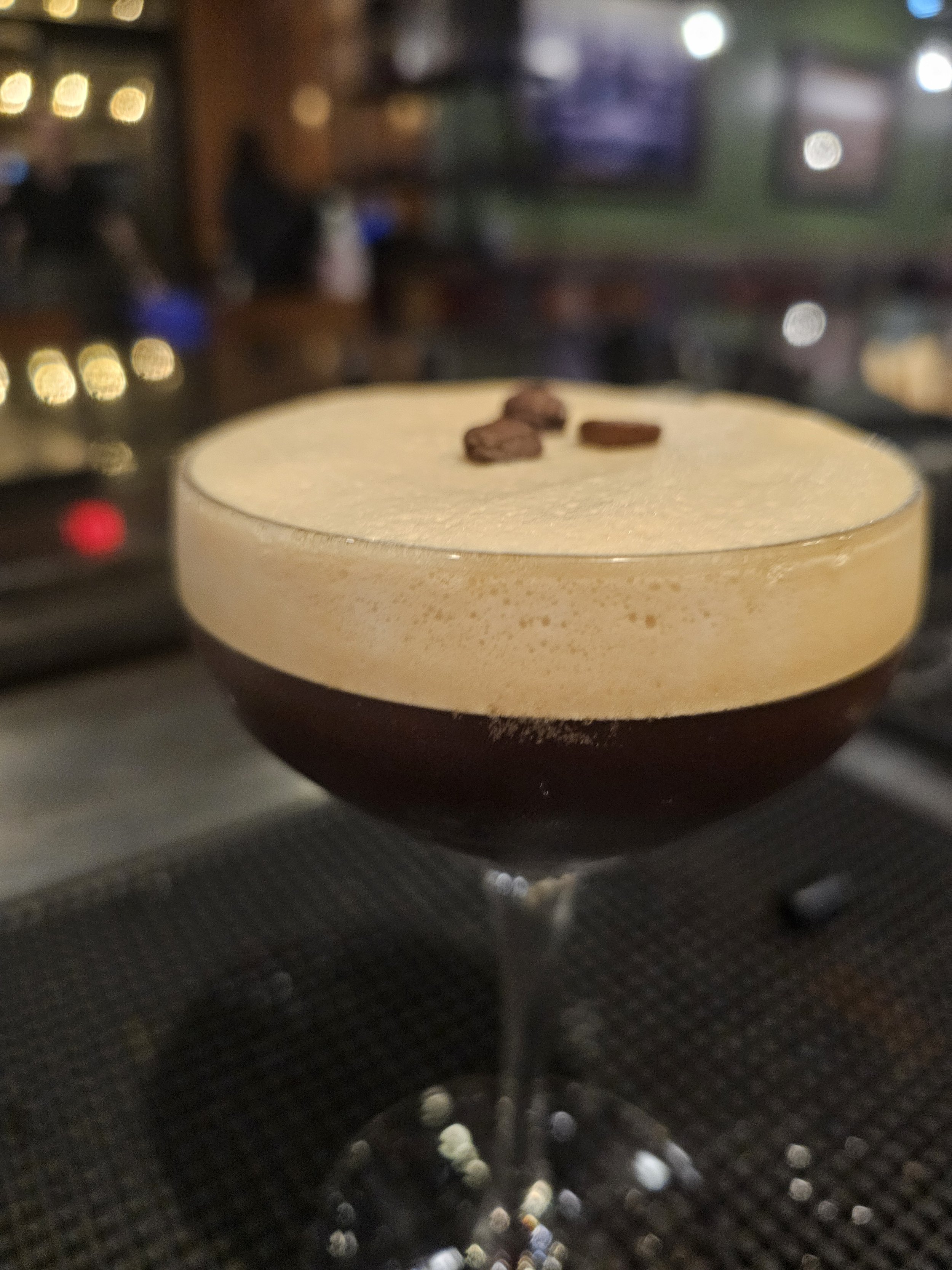 A close-up of a cocktail in a coupe glass with a dark bottom and a creamy frothy top, decorated with a few oat flakes, in a dimly lit bar or restaurant setting.