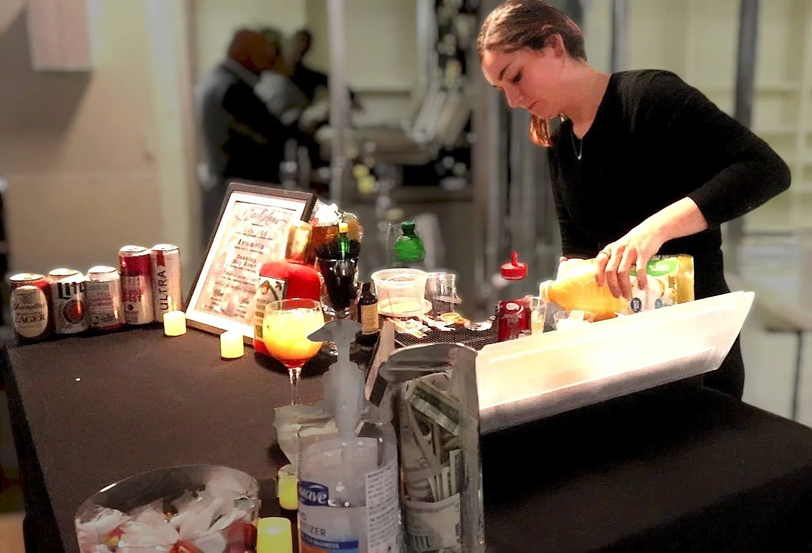 A woman preparing food at a station with various condiments, bottles, and beverages, with candles and a drink on the table, and another person blurred in the background.