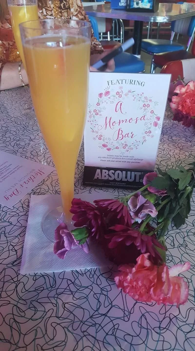 A tall glass of orange mimosa on a table decorated with pink and purple flowers, an 'A Momosa Bar' sign, and a patterned tablecloth at a celebration or event.