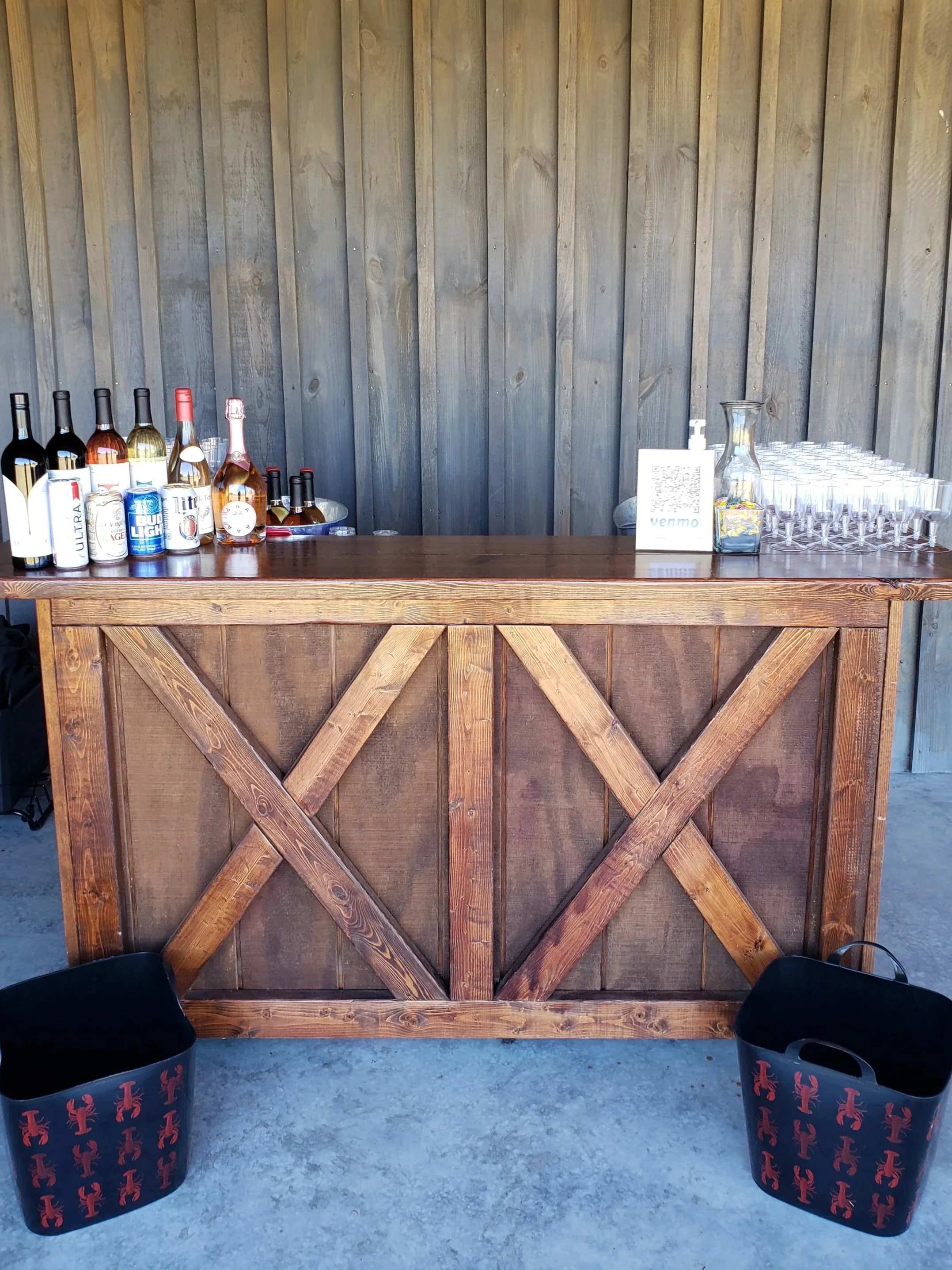 A wooden bar with bottles of liquor, a QR code sign, and glasses, set against a wood-paneled wall, with two black baskets decorated with red lobsters in front.