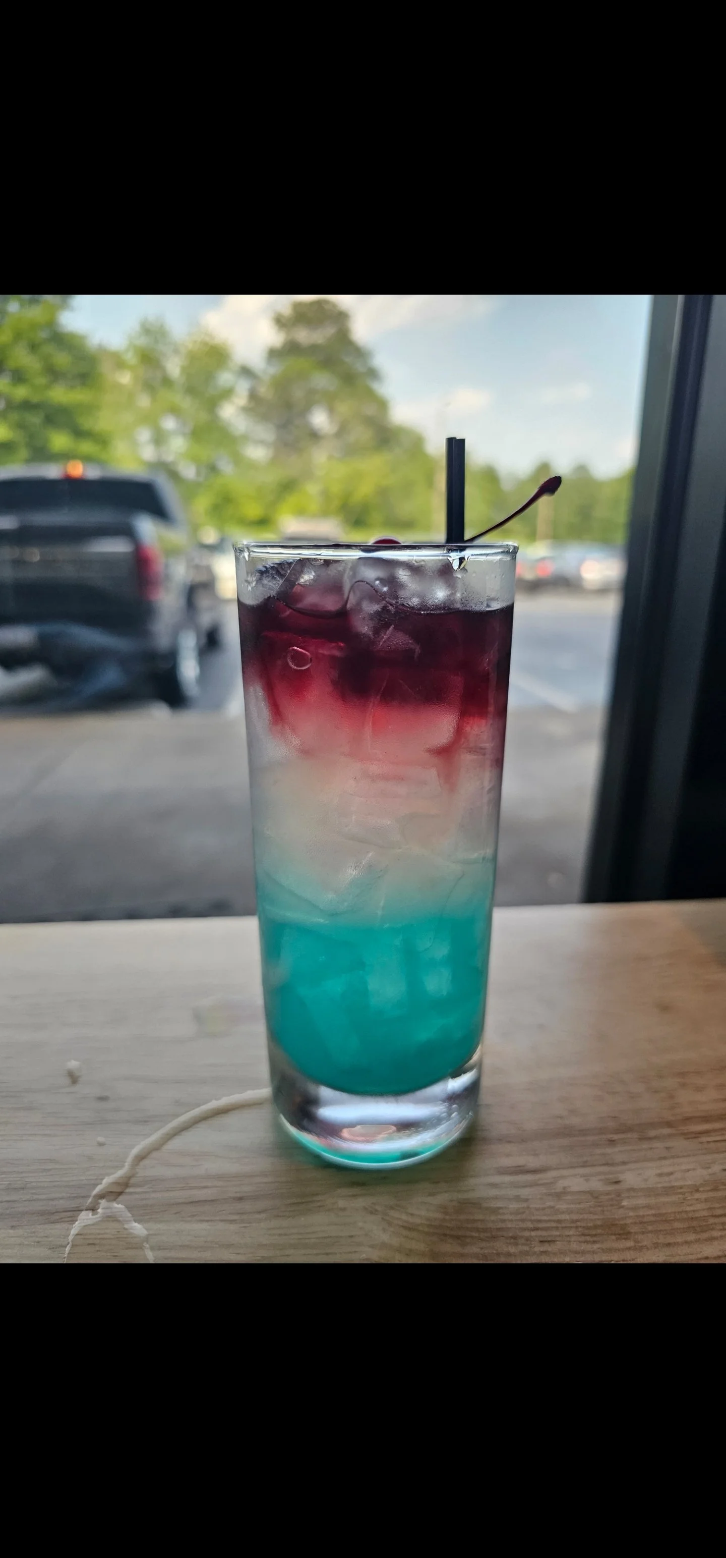 A multi-colored layered cocktail with ice, served in a tall glass with a straw and a cherry on top, on a wooden table with a window showing trees and parked cars in the background.