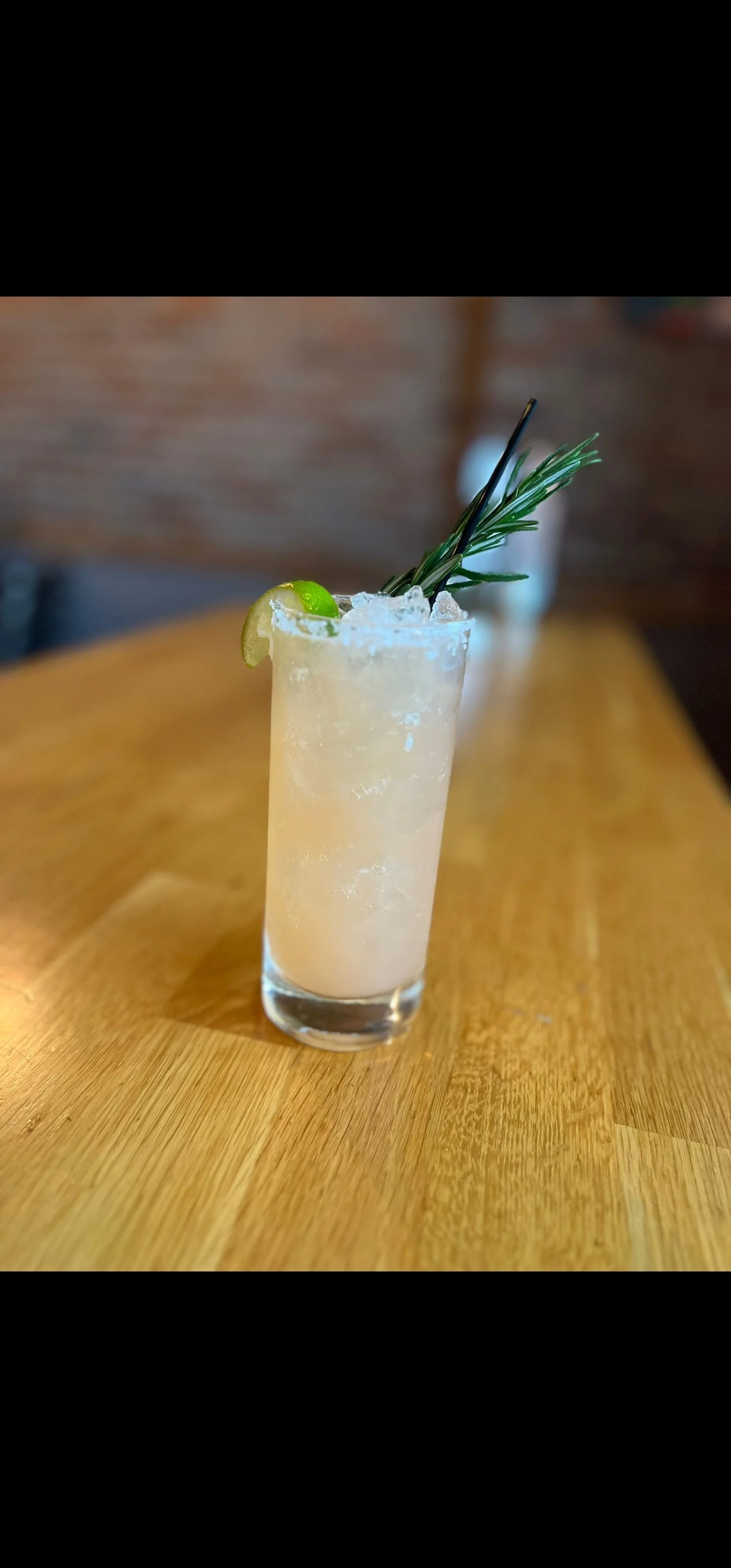 A tall glass filled with a pale, frothy cocktail and crushed ice, garnished with a lime wedge, a sprig of rosemary, and a black cocktail pick, on a wooden table.