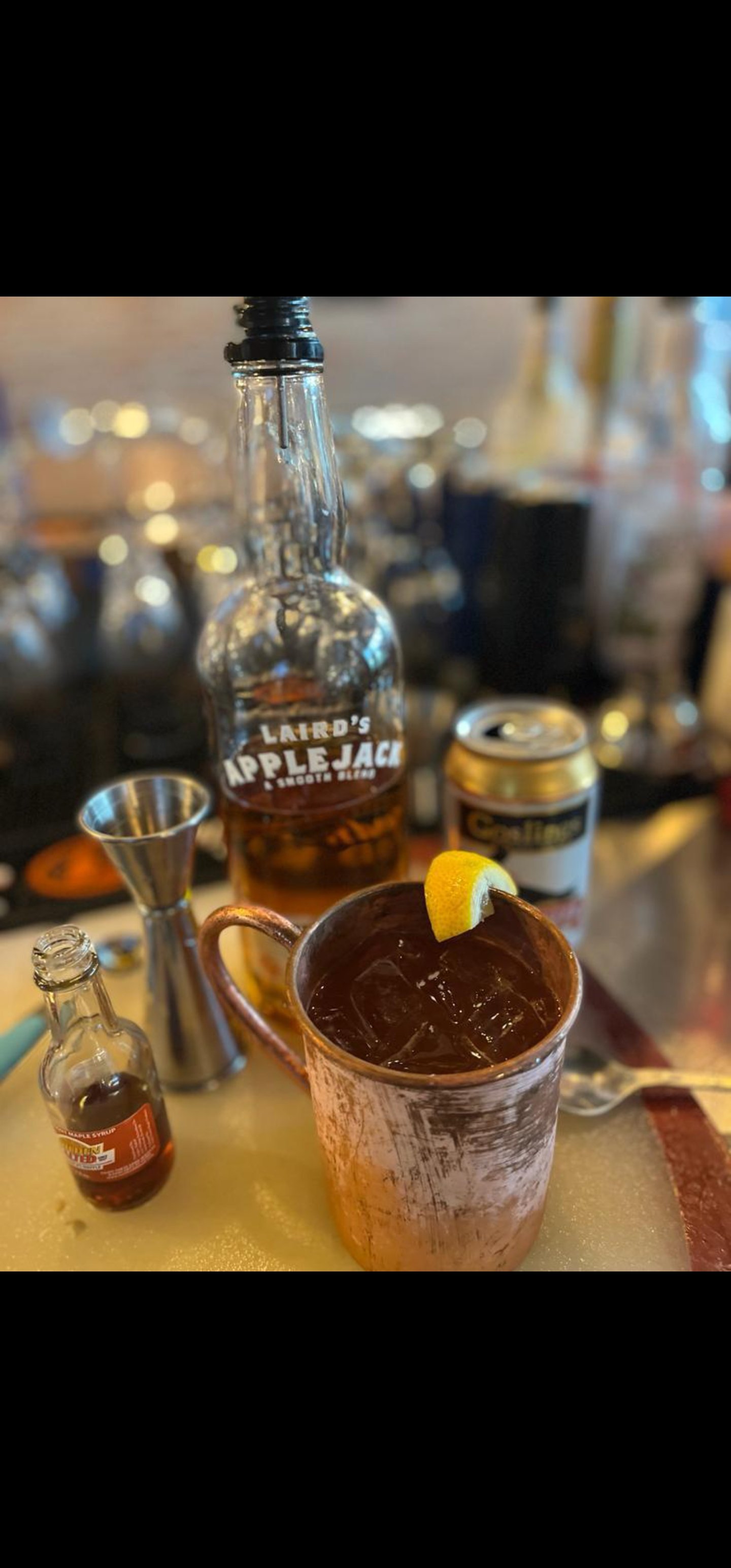 A copper mug filled with dark iced beverage and ice cubes, garnished with a lemon wedge, on a yellow cutting board with bar tools and alcohol bottles in the background.