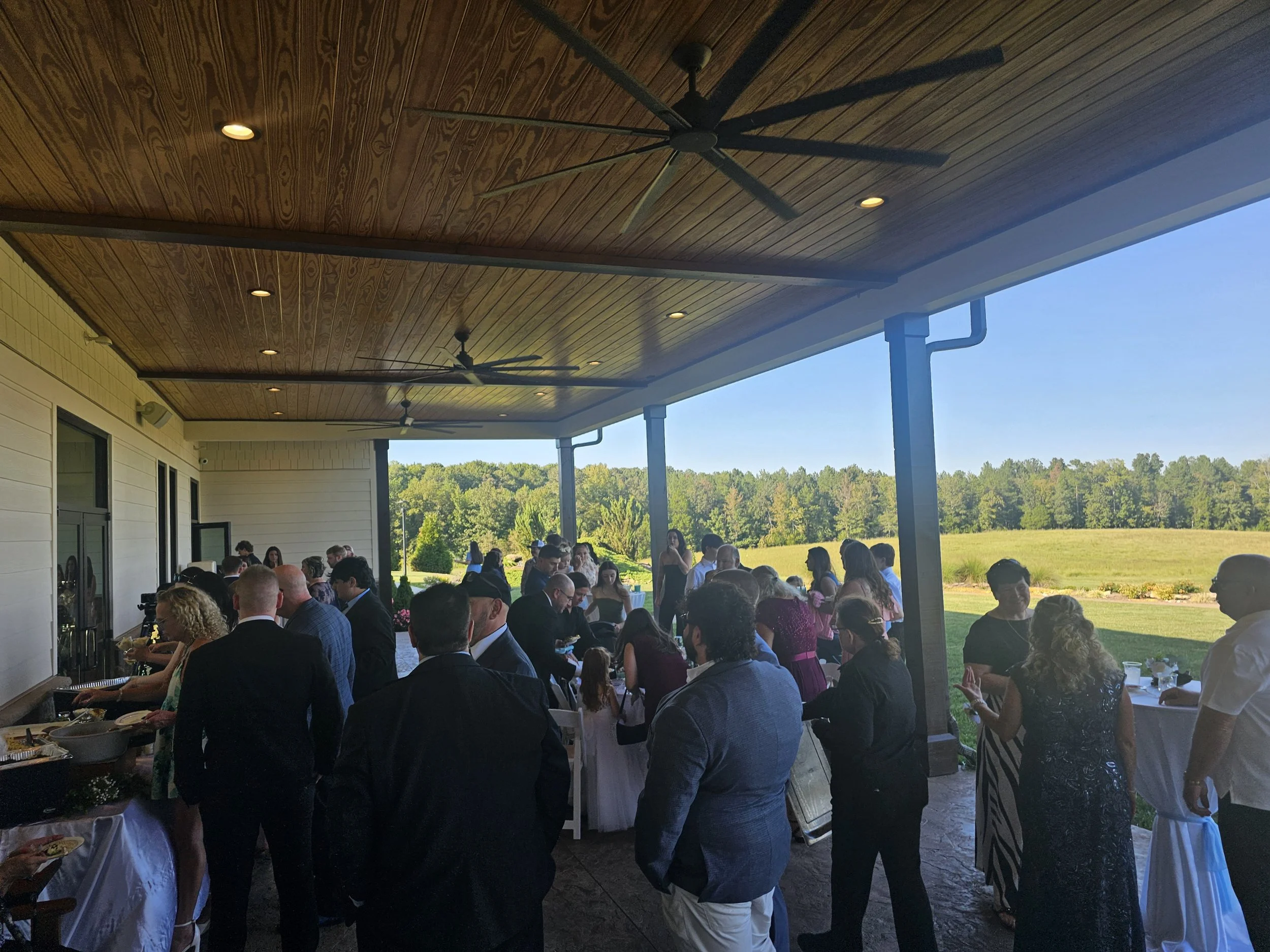 People gathered at an outdoor event with a scenic view of a green field and trees, under a covered patio with a wooden ceiling and ceiling fans.