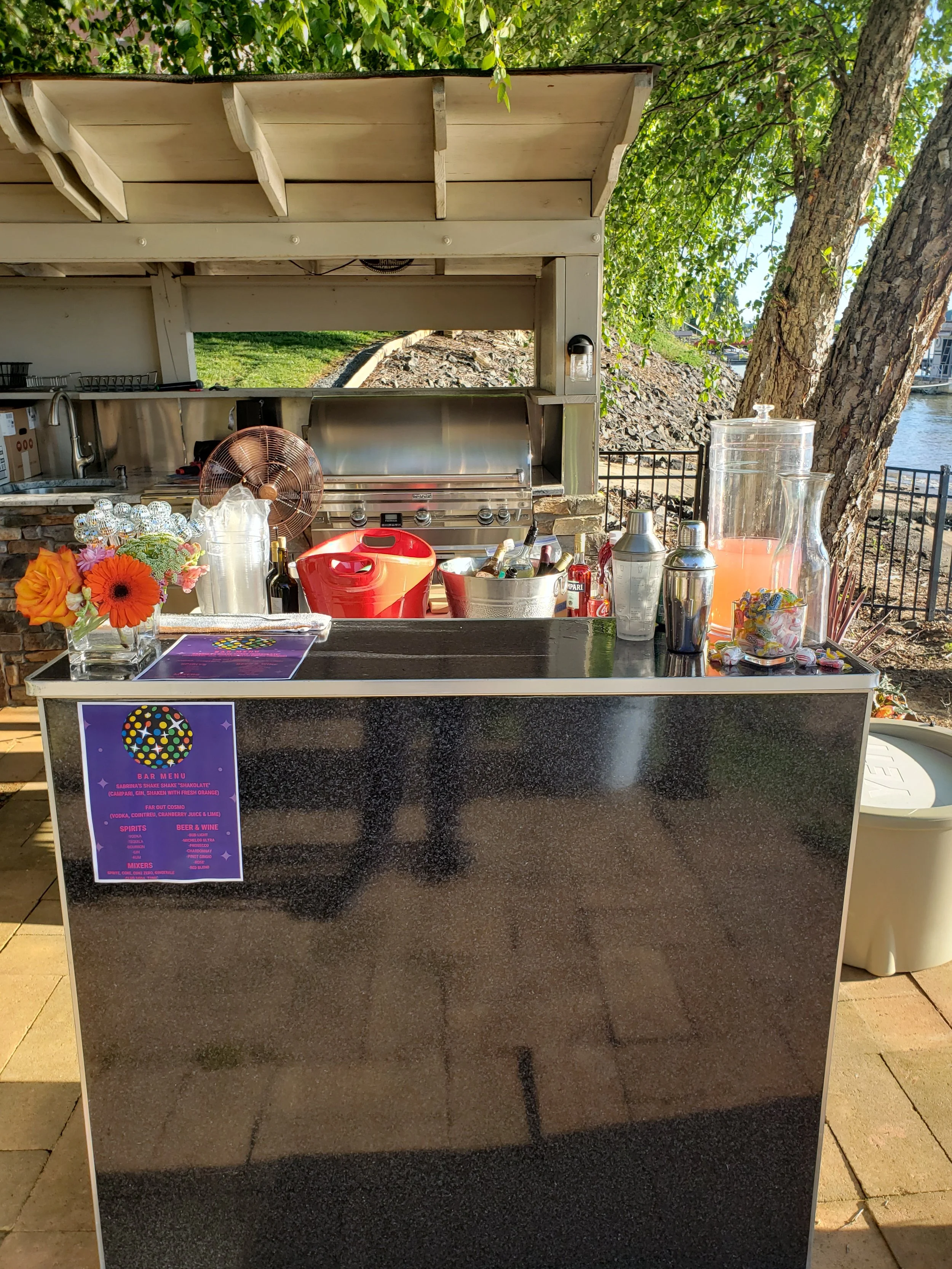 An outdoor bar setup with a black counter, a large glass beverage dispenser, and various bottles and bar tools. In the background, there is a barbecue grill, a paper fan, a flower arrangement, and a bright sunny setting with trees and water nearby.