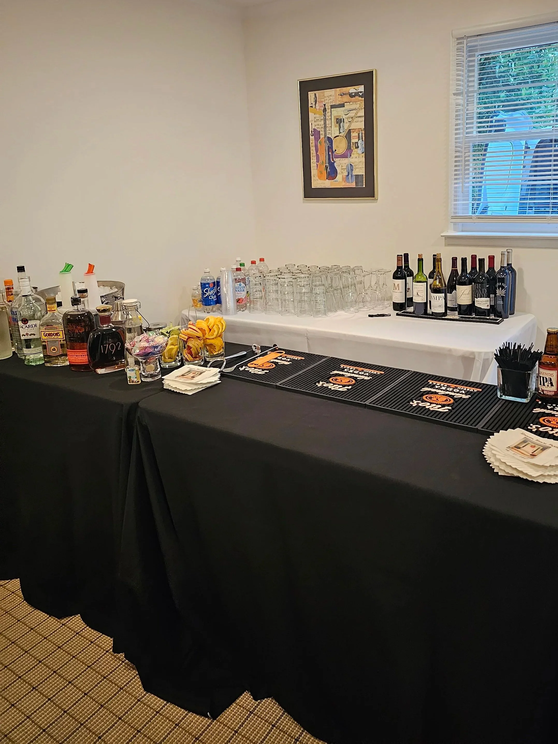 A black table with alcohol bottles, glassware, napkins, and bar accessories, set up for a party in a room with a framed artwork on the wall and blinds covering a window.